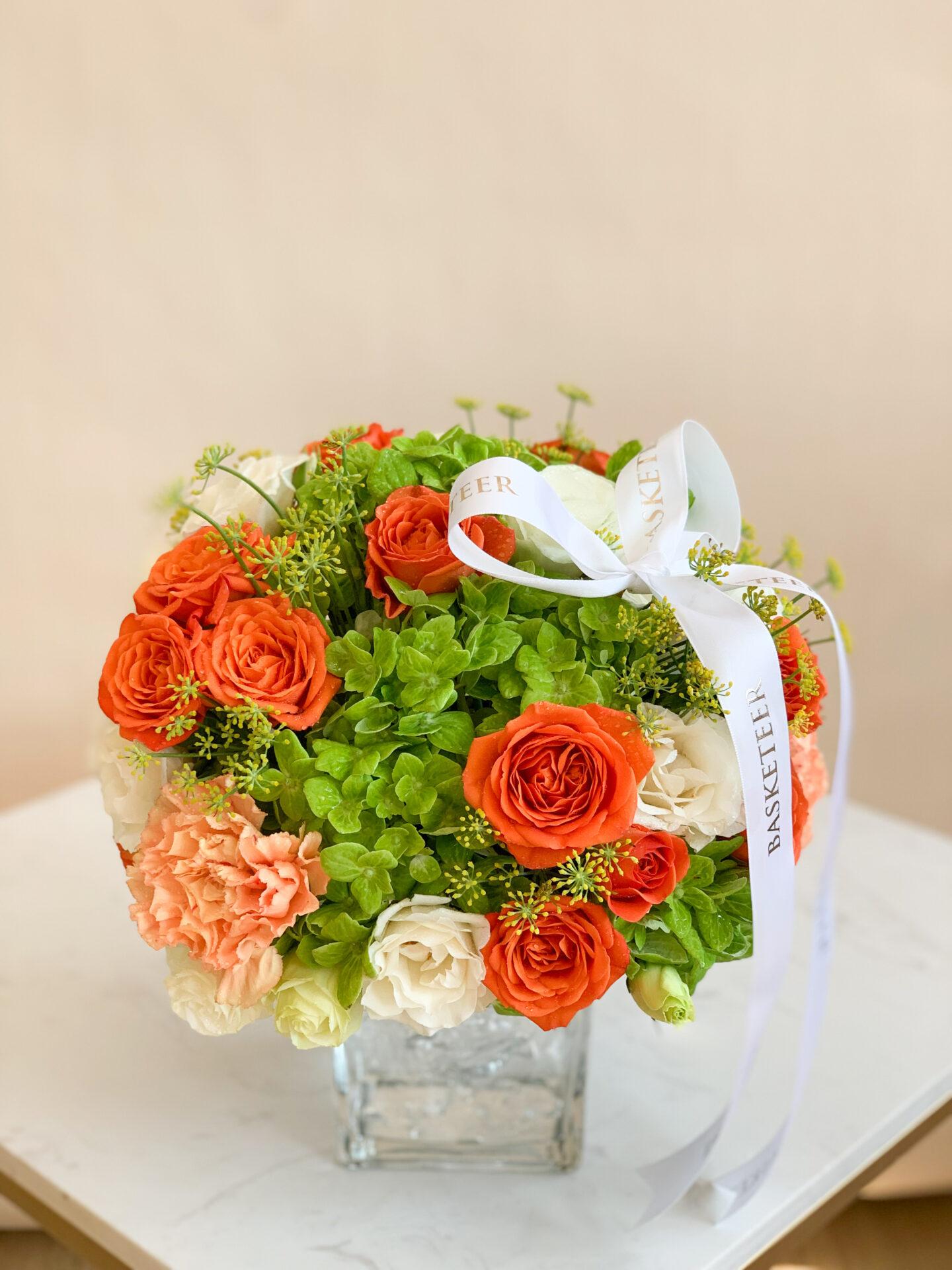 A lavish floral arrangement featuring radiant orange and pristine white roses, lush green hydrangeas, and elegant greenery graces a beautifully designed glass vase. This centerpiece rests atop a table covered in an exquisite beige cloth with a sumptuous gray satin runner flowing across it. Enhancing the opulent ambiance, the table is adorned with vibrant oranges, clusters of juicy grapes, artfully halved orange slices, two refined white candles casting a gentle glow, and thoughtfully placed books. The scene exudes luxury and sophistication, punctuated by glimpses of the Basketeer logo on select accents that personalize this enchanting tableau.