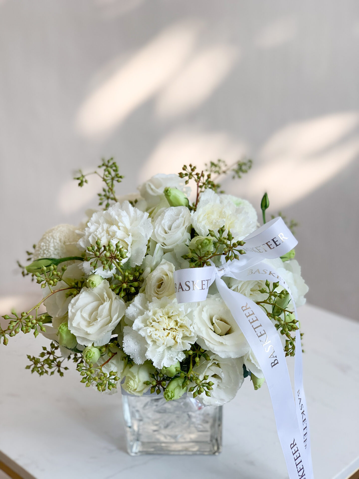 A luxurious bouquet of pristine white roses and lush green hydrangeas is elegantly arranged in a crystal-clear glass vase, gracing a stack of two sophisticated books on a tastefully round table adorned with an exquisite beige tablecloth. A delicate white cloth drapes beside the vase, adding to the refined ambiance. Nearby, a plate brimming with fresh green grapes and a succulent pear tempts the senses. Two ivory spiral candles in glass holders cast a warm, inviting glow over the scene. The finishing touch: 