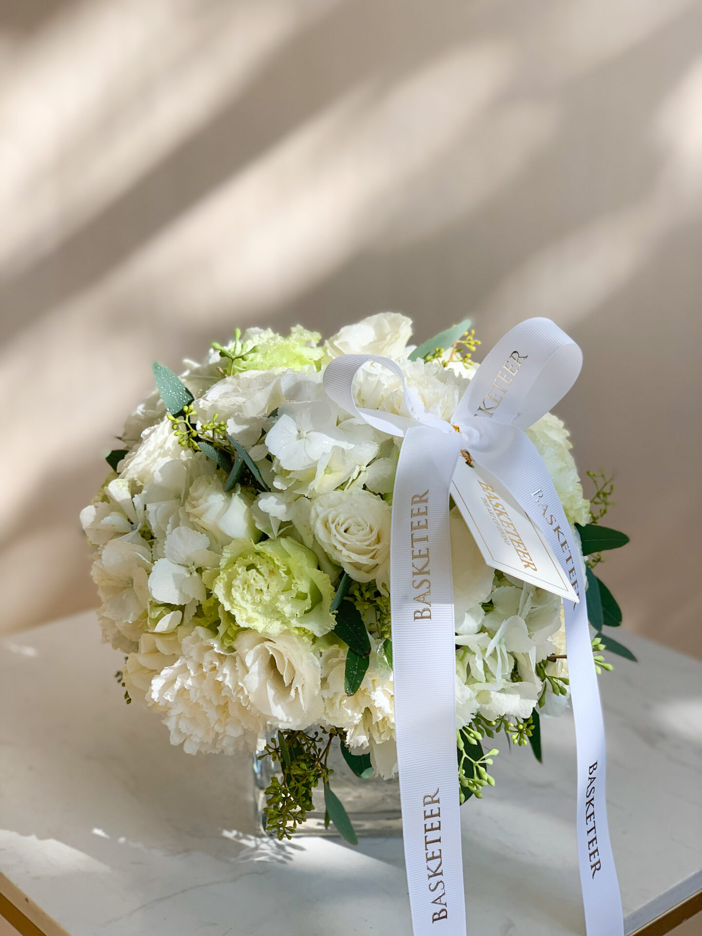 A round table, epitomizing luxury and grace, is exquisitely adorned with a Basketeer signature touch. At its center, a crystal-clear glass vase brimming with pristine white roses and lush greenery exudes elegance. Flanking the vase are two sophisticated white taper candles in sleek glass holders, casting a serene glow over the scene. A small cluster of fresh green grapes adds an inviting charm. The table is finished with a beige cloth artfully draped over its edge, hinting at hidden literary treasures underneath. The word 