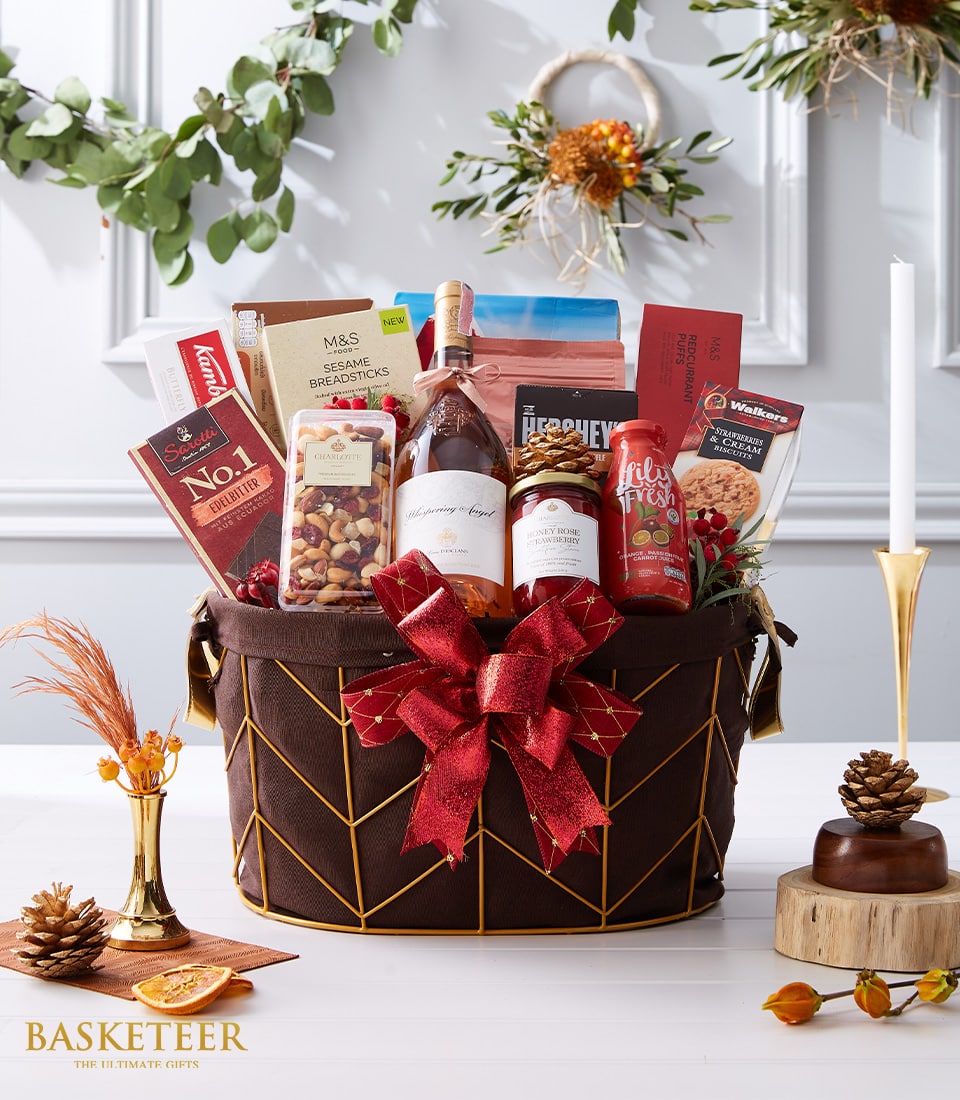 Festive brown-gold basket with rosé wine, premium chocolates, mixed nuts, breadsticks, jam, juice, biscuits, and red bow
