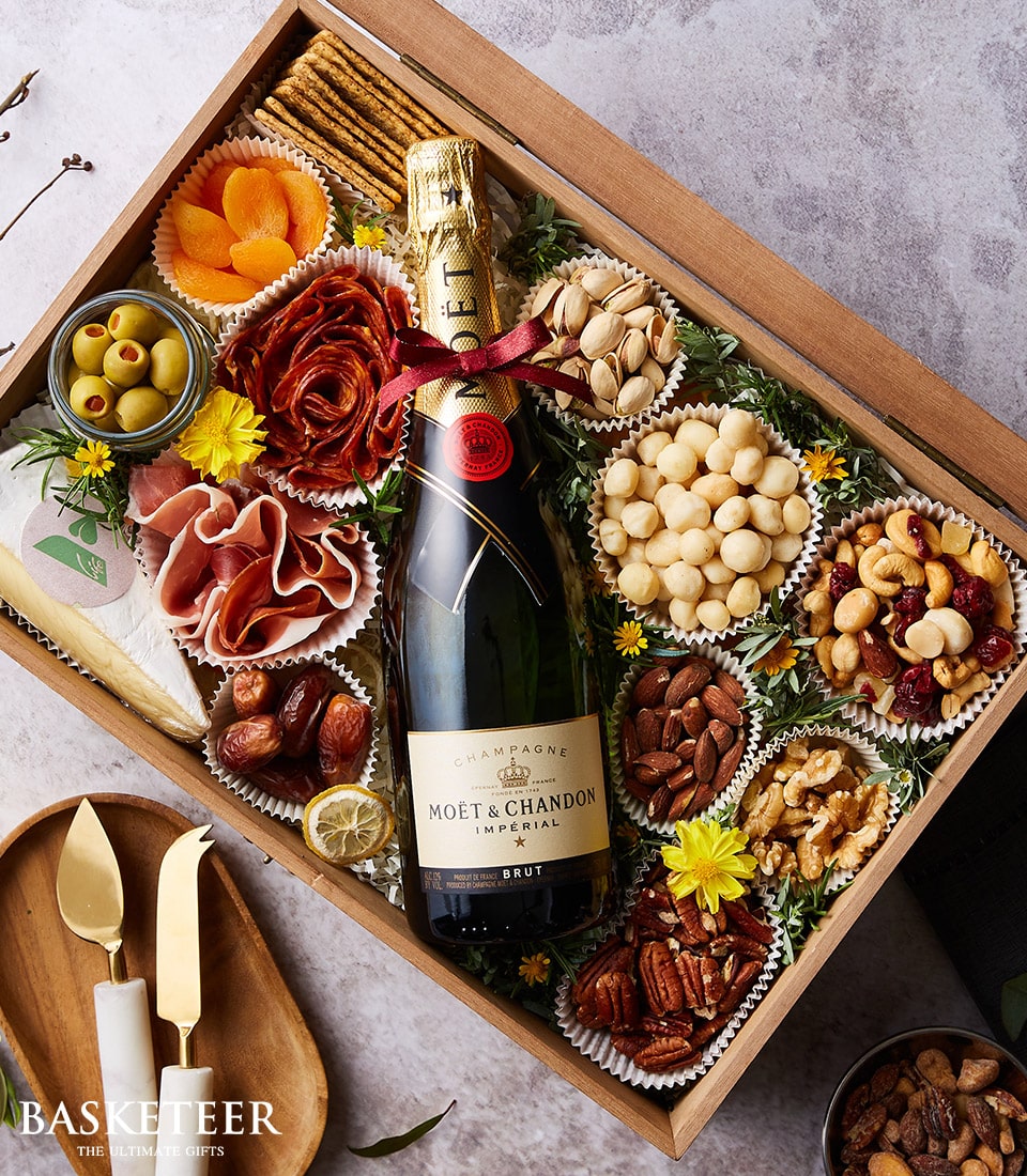 Experience the epitome of luxury with this exquisite Basketeer gift box. A bottle of Moët & Chandon champagne reigns supreme, complemented by a curated selection of gourmet delights including artisan nuts, fine cheeses, succulent dried fruits, savory olives, and premium charcuterie. The scene is enhanced by two elegant cheese knives resting beside the opulent arrangement. This lavish presentation is adorned with decorative flowers and proudly features the distinguished Basketeer logo on a refined ribbon and box, inviting you to indulge in a moment of pure sophistication.