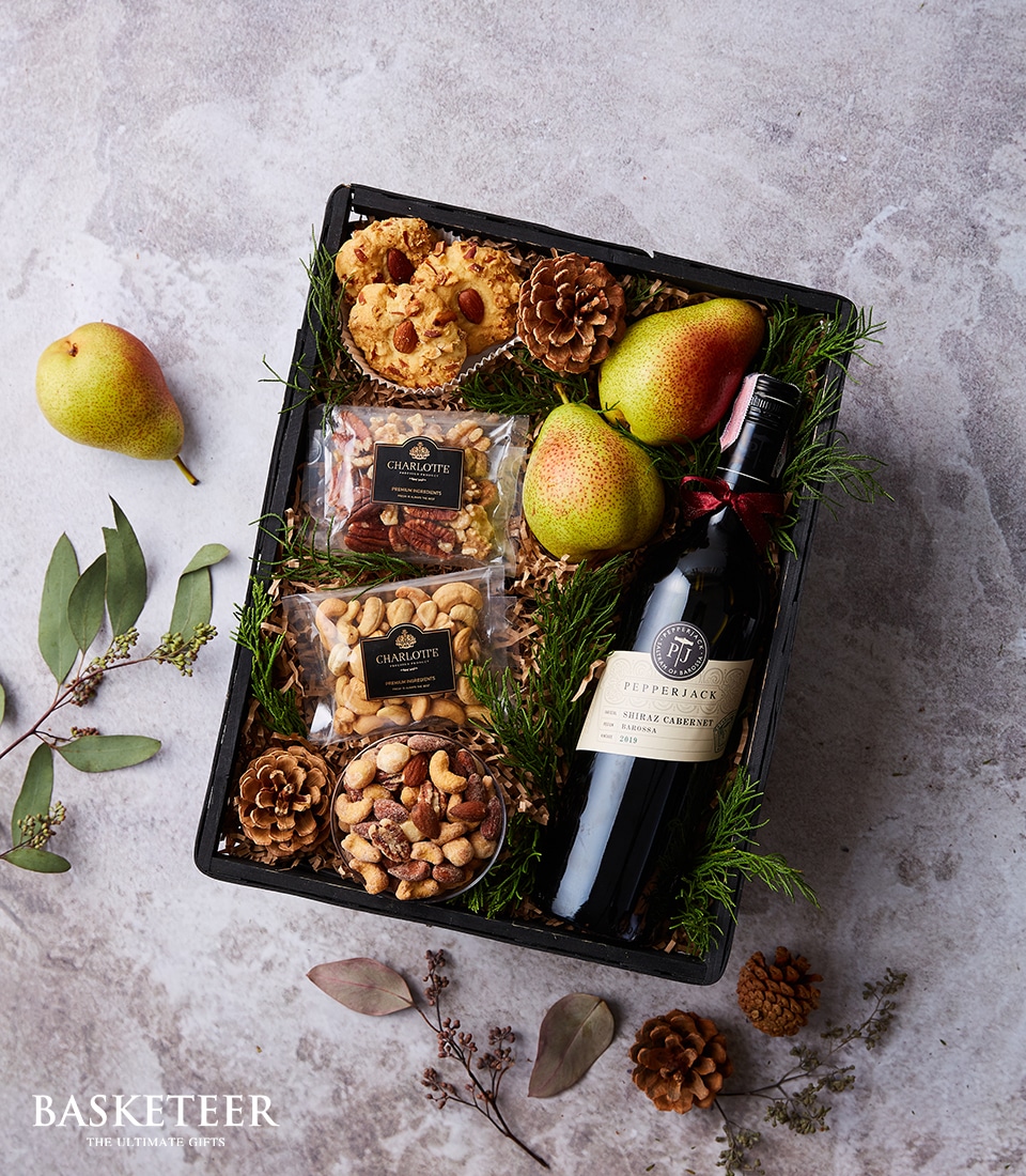 A luxurious Basketeer gift basket is artfully arranged on a sophisticated gray surface, surrounded by scattered leaves and rustic pinecones. Nestled within the elegant basket, branded with the Basketeer logo, you’ll find a bottle of Pepperjack Shiraz Cabernet wine inviting you to indulge in its rich flavors. Accompanying this fine wine are several perfectly ripened pears, two packets of gourmet snacks that promise delightful culinary experiences, and two bags of premium mixed nuts for an added touch of sophistication. A charming small pinecone and vibrant leafy greens complete this opulent display, making it an irresistibly inviting gift for any occasion.