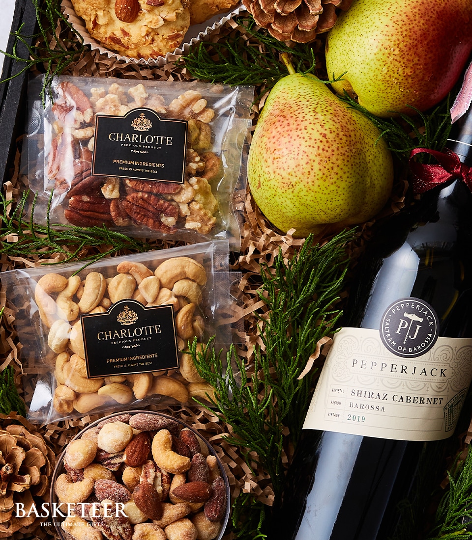 A lavish gourmet gift basket curated by Basketeer, featuring a rich assortment of premium nuts, an exquisite box of decadent holiday sweets adorned with the elegant Basketeer ribbon, two perfectly ripened fresh pears, and a distinguished bottle of Pepperjack Shiraz Cabernet wine from Barossa Valley, vintage 2019. The luxurious contents are meticulously arranged on a plush bed of shredded brown paper and complemented by festive pinecones for an inviting touch.