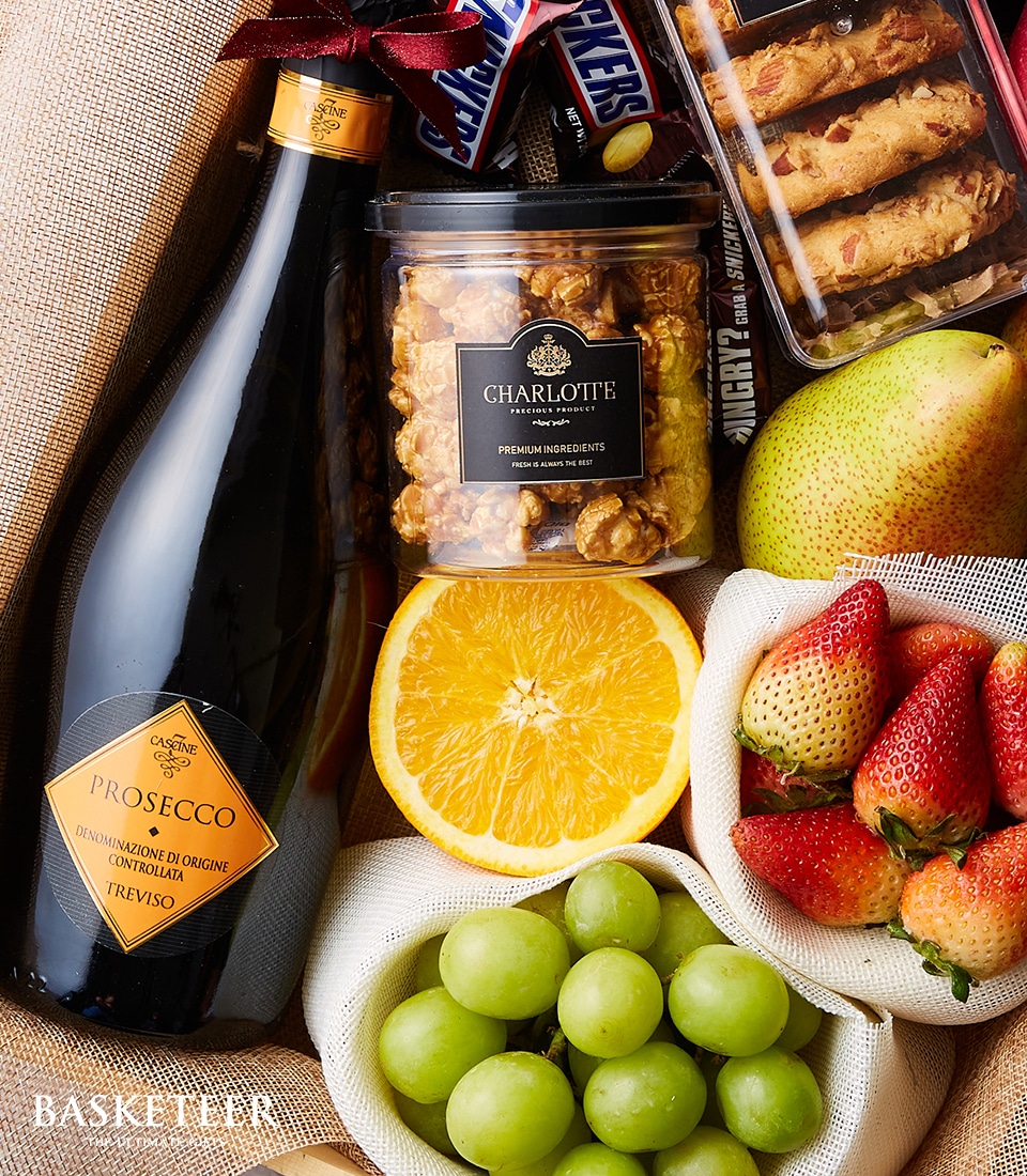 A lavish gift basket exuding opulence and inviting indulgence, featuring a premium bottle of Prosecco that beckons celebration. Nestled alongside is a luxurious tub of caramel popcorn, assorted gourmet cookies, and an artisanal Snickers bar. Fresh fruits including a pear, a halved vibrant orange, succulent strawberries, and crisp green grapes add a touch of natural elegance. This curated collection is elegantly presented with the Basketeer logo proudly displayed on the ribbon and labeled box, guaranteeing an unforgettable gifting experience.