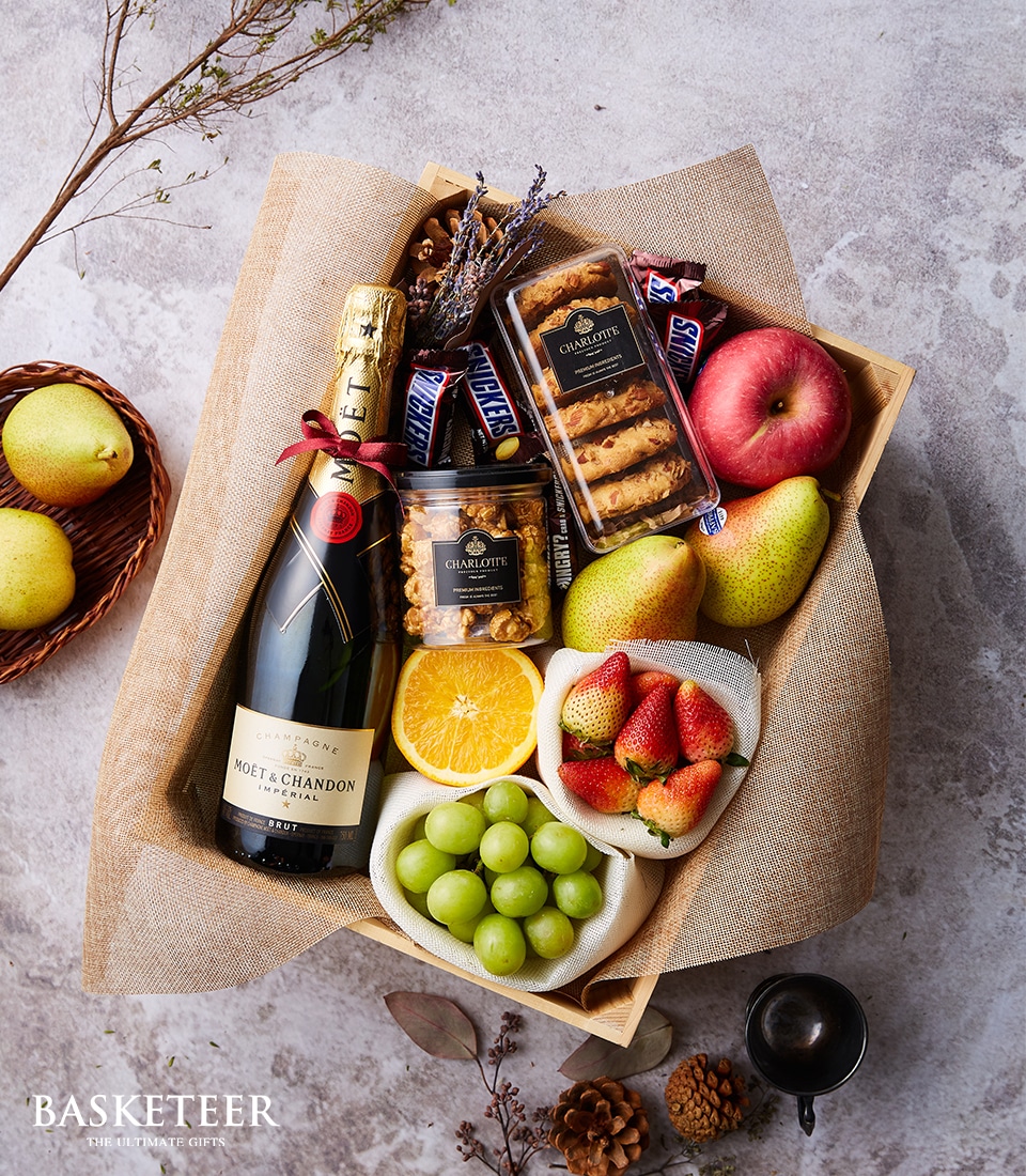 An exquisite gift box by Basketeer, featuring a bottle of Moët & Chandon champagne nestled amidst an array of indulgent treats. The selection includes decadent assorted chocolates, an elegant jar of honey, fresh strawberries, vibrant green grapes, a zesty orange slice, a crisp red apple, two juicy pears, and a fragrant pack of dried lavender. All items are artfully arranged on a rustic backdrop for an inviting presentation. The luxurious scene is completed with the Basketeer branding gracefully displayed in the bottom left corner.