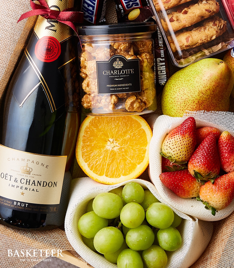 An elegant and luxurious gift basket from Basketeer, adorned with their signature ribbon, showcasing a bottle of Moët & Chandon champagne as its centerpiece. Surrounding it is a delightful selection of Charlotte's gourmet caramel popcorn in an exquisite tub, ripe fresh strawberries, succulent green grapes, a perfectly halved orange radiating citrusy freshness, and a crisp pear. Premium chocolate bars and an artisanal pack of cookies complete this opulent ensemble. All the items are meticulously arranged in a finely woven basket that exudes sophistication and invites indulgence.