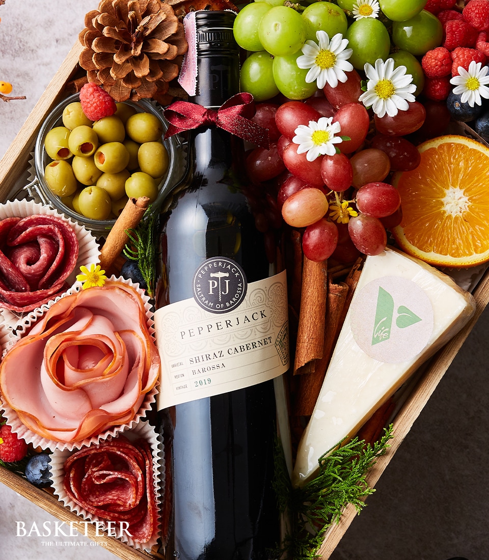 A beautifully curated wooden gift basket exudes luxury, featuring a bottle of Pepperjack Shiraz Cabernet wine. This elegant ensemble is adorned with an array of fresh fruits including plump grapes and a vibrant orange slice, complemented by an assortment of green and black olives. A premium block of cheese and delicately arranged cured meats fashioned into intricate floral shapes add to the opulence. The presentation is artfully garnished with fresh herbs and flowers, inviting indulgence. Enhancing the allure, a ribbon embellished with the Basketeer logo subtly drapes over the arrangement, adding a touch of branded sophistication to this exquisite offering from Basketeer.