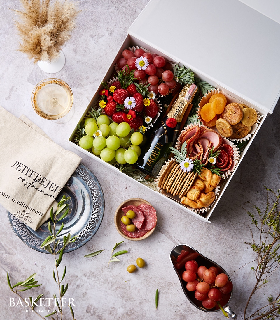 A luxurious Basketeer gift box lavishly arranged with an assortment of fresh fruits, gourmet dried apricots, artisan crackers, premium cheese, and savory salami, alongside an exquisite bottle of Moët champagne. The scene is elegantly set with a glass of champagne, a pristine linen napkin, a sophisticated blue plate heaped with more delectable treats, and a small bowl of olives—all meticulously placed upon an opulent marble surface. A ribbon featuring the Basketeer logo adds a touch of brand elegance to this indulgent arrangement.
