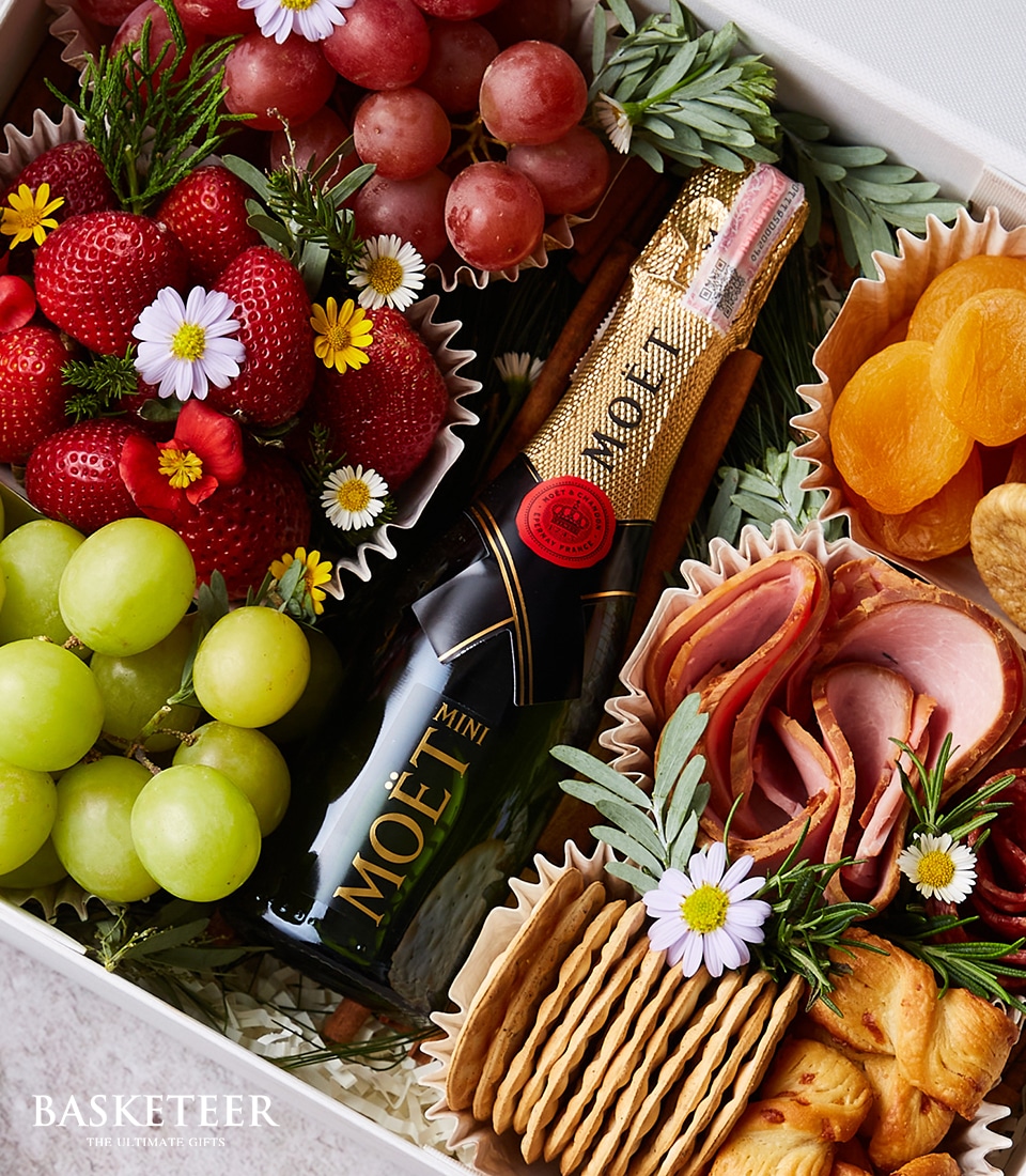 A luxurious gift box beautifully arranged with a mini bottle of Moët champagne, accompanied by fresh strawberries, succulent red and green grapes, delectable dried apricots, an assortment of gourmet cold cuts, savory crackers, and crispy pastry twists. The presentation is enhanced by delicate small white flowers adding a touch of elegance. The sophisticated scene is accented with the prestigious 