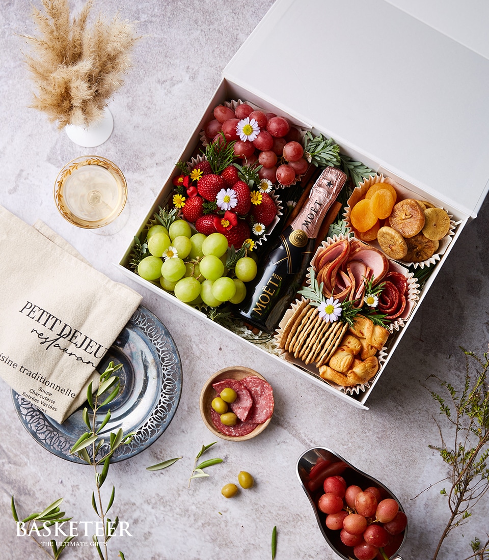 A luxurious gourmet gift box from Basketeer, elegantly branded with the Basketeer logo, brims with a premium selection of fresh fruits, artisan crackers, succulent dried apricots, expertly cured meats, and decadent cheeses. Highlighting the sumptuous assortment is a prestigious bottle of Moët & Chandon champagne. The scene is impeccably set on a stylishly arranged table adorned with a delicate ribbon bearing the Basketeer emblem. A crystal glass filled with sparkling champagne stands ready alongside an exquisite napkin inscribed 