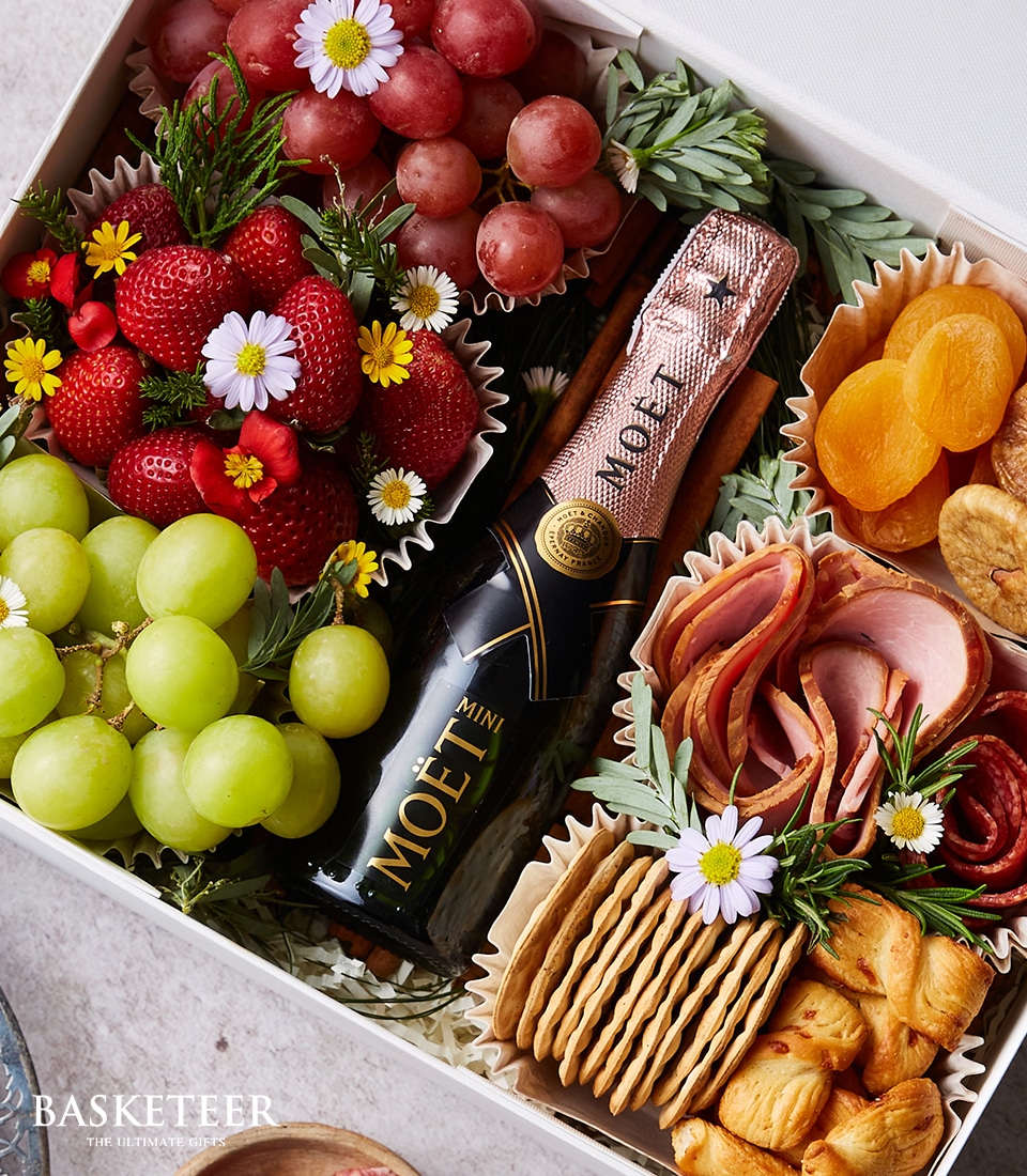 An exquisitely curated gift basket exudes luxury and elegance, featuring a petite bottle of Moët champagne nestled among vibrant clusters of red and green grapes, plump strawberries, a selection of premium dried fruits, artisanal crackers, gourmet meats, and fine cheeses. This sumptuous arrangement is tastefully garnished with delicate white flowers and lush greenery. The prestigious 