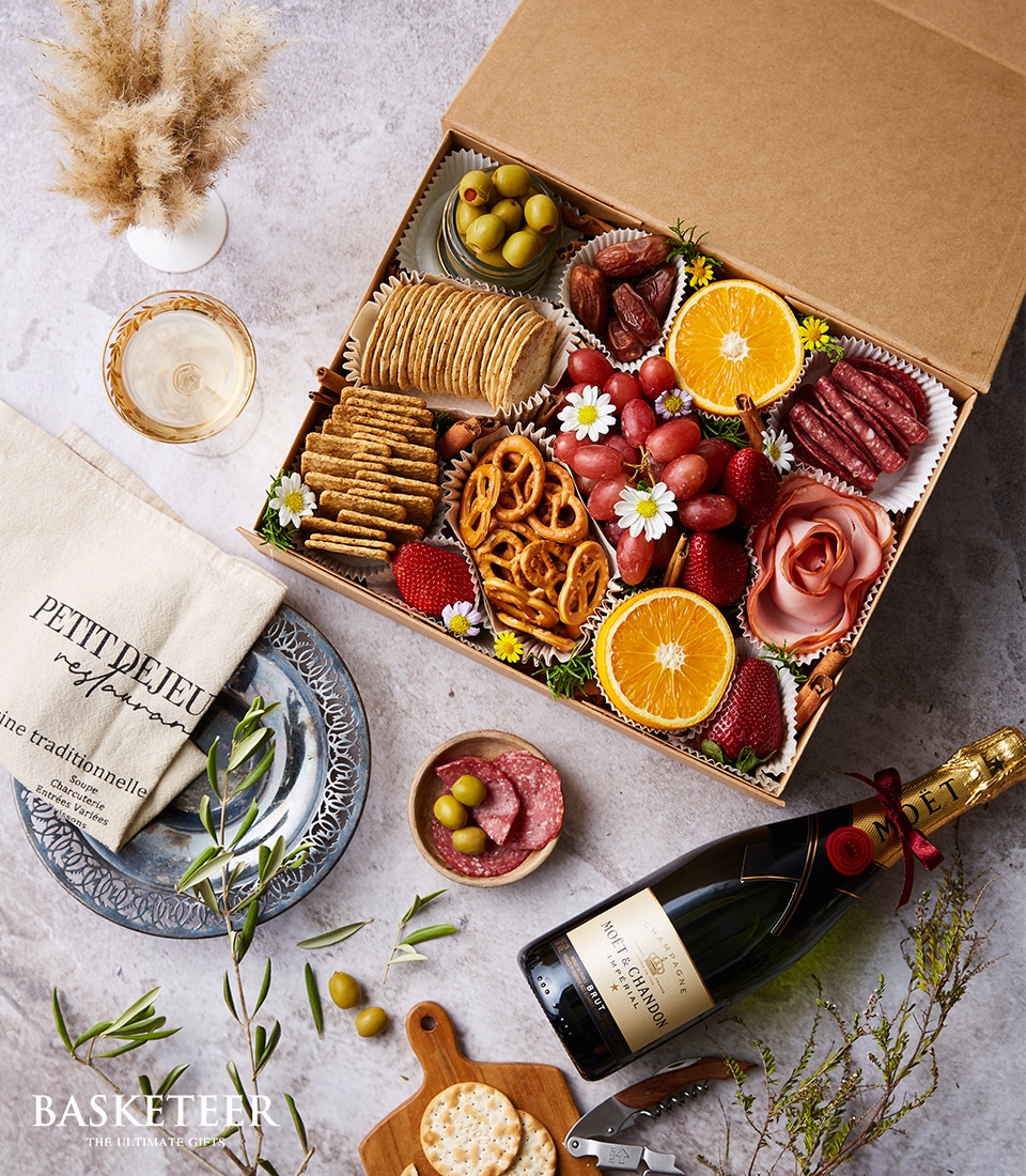 A lavish charcuterie gift box brimming with sumptuous sliced oranges, plump grapes, marinated olives, and ripe strawberries. Delight in an assortment of gourmet meats, crisp pretzels, and artisan crackers. The scene is completed with a freshly baked baguette resting beside a creamy cheese selection and a small bowl of delectable olives. Elegantly presented alongside is a bottle of Peller Estates champagne for moments of indulgence, atop a pristine linen napkin accentuated by delicate decorative flowers. A ribbon adorned with the Basketeer logo befits this luxurious ensemble, epitomizing sophistication and opulence.  