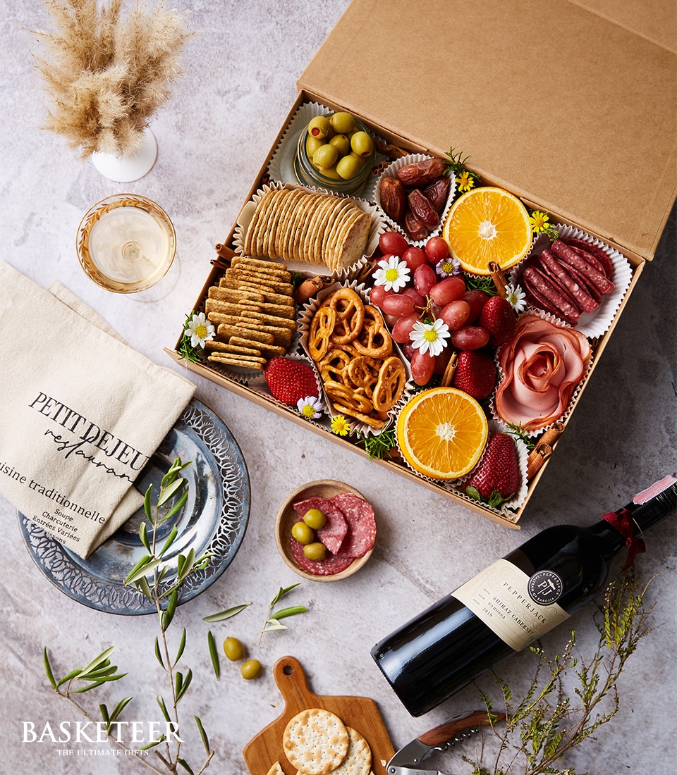 Alt text: An exquisite charcuterie box by Basketeer, elegantly arranged with succulent sliced oranges, plump grapes, juicy strawberries, savory olives, crispy pretzels, gourmet salami, and artisanal crackers. Encircling this luxurious assortment is a chic bag labeled 