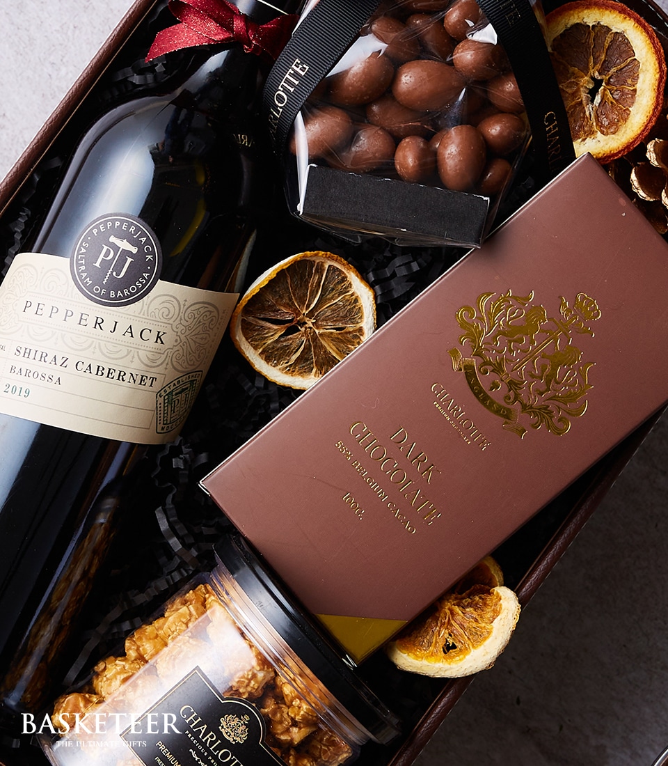 An elegantly curated gourmet gift basket perfect for indulgence. Nestled within a handwoven basket, you'll find a distinguished bottle of Pepperjack Shiraz Cabernet wine ready to tantalize the taste buds. Alongside, discover a sophisticated box of Charlotte dark chocolate, an artisan jar of caramel popcorn, and a luxurious container of chocolate-covered nuts. To complete this sensory delight, there are delicately dried orange slices adding a touch of zest. The scene is accented with the esteemed 