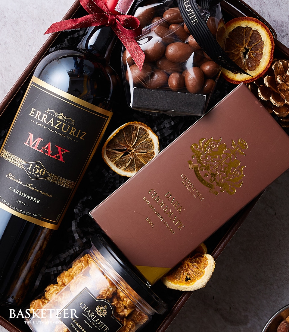 An elegantly curated gift basket features a sumptuous bottle of Errazuriz Max wine, a sophisticated box of Charlotte dark chocolate, an indulgent container of chocolate-covered nuts, an exquisite jar of caramel popcorn, and artfully arranged dried orange slices. These luxurious treats are nestled together in a festive display. The presentation is elevated with the Basketeer logo prominently displayed on a stylish ribbon and box, ensuring this gift exudes opulence and invites delight.