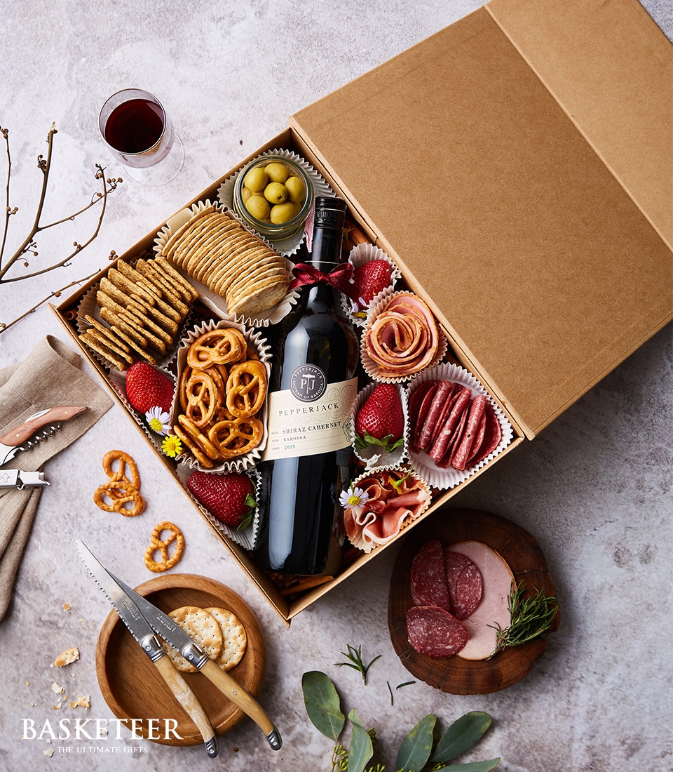 A meticulously curated charcuterie box by Basketeer, featuring an exquisite selection of savory deli meats, artisanal cheeses, luscious strawberries, briny olives, and crispy pretzels complemented by gourmet crackers. Nestled within the arrangement is a distinguished bottle of red wine and a half-filled wine glass invitingly placed beside it. Accompanying this luxurious spread is a charming wooden board with knife and sliced sausage elegantly presented for your tasting pleasure. The elegant box bears the prestigious 