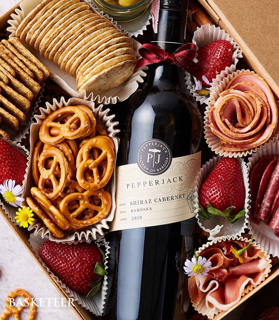 Indulge in the decadent offerings of a Basketeer gift box, featuring a sophisticated bottle of Pepperjack Shiraz Cabernet wine. This luxurious ensemble is artfully arranged with gourmet treats including elegantly wrapped pretzels, succulent sliced strawberries, savory rolled meats, and an exquisite assortment of artisanal crackers. Each element is thoughtfully presented in refined cupcake liners, creating a visually inviting and sumptuous display. Completing this elegant presentation is the distinguished Basketeer logo gracing the bottom left corner, signifying quality and elegance.