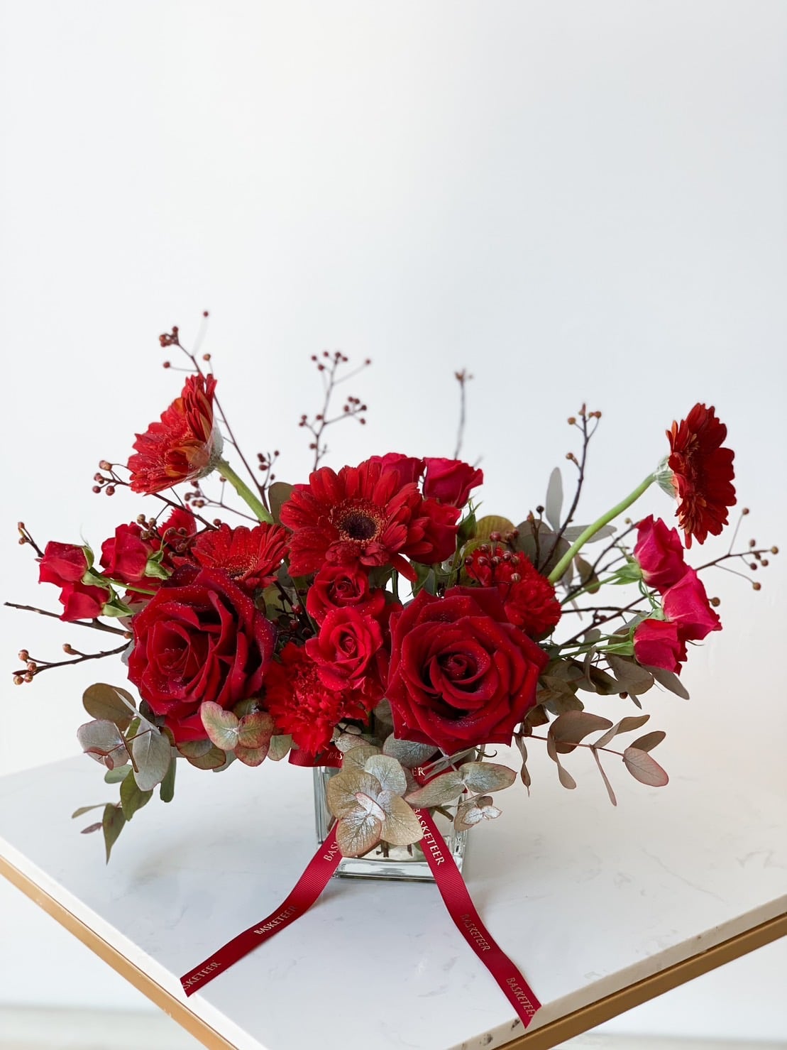 A vibrant floral arrangement with an assortment of red roses, gerbera daisies, and other red flowers, accented with greenery and eucalyptus leaves. The bouquet, displayed in a clear vase with red ribbons, rests on a white marble surface against a plain background.