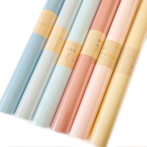 Six elegant rolls of pastel-colored wrapping paper stand gracefully upright. The luxurious hues include light blue, light green, light pink, darker pink, pale yellow, and cream. Each sophisticated roll is adorned with a beige band featuring intricate text and symbols, effortlessly elevating their charm. A tastefully positioned box with the Basketeer logo completes this exquisite presentation, inviting you to add a touch of elegance to any gift-giving occasion.