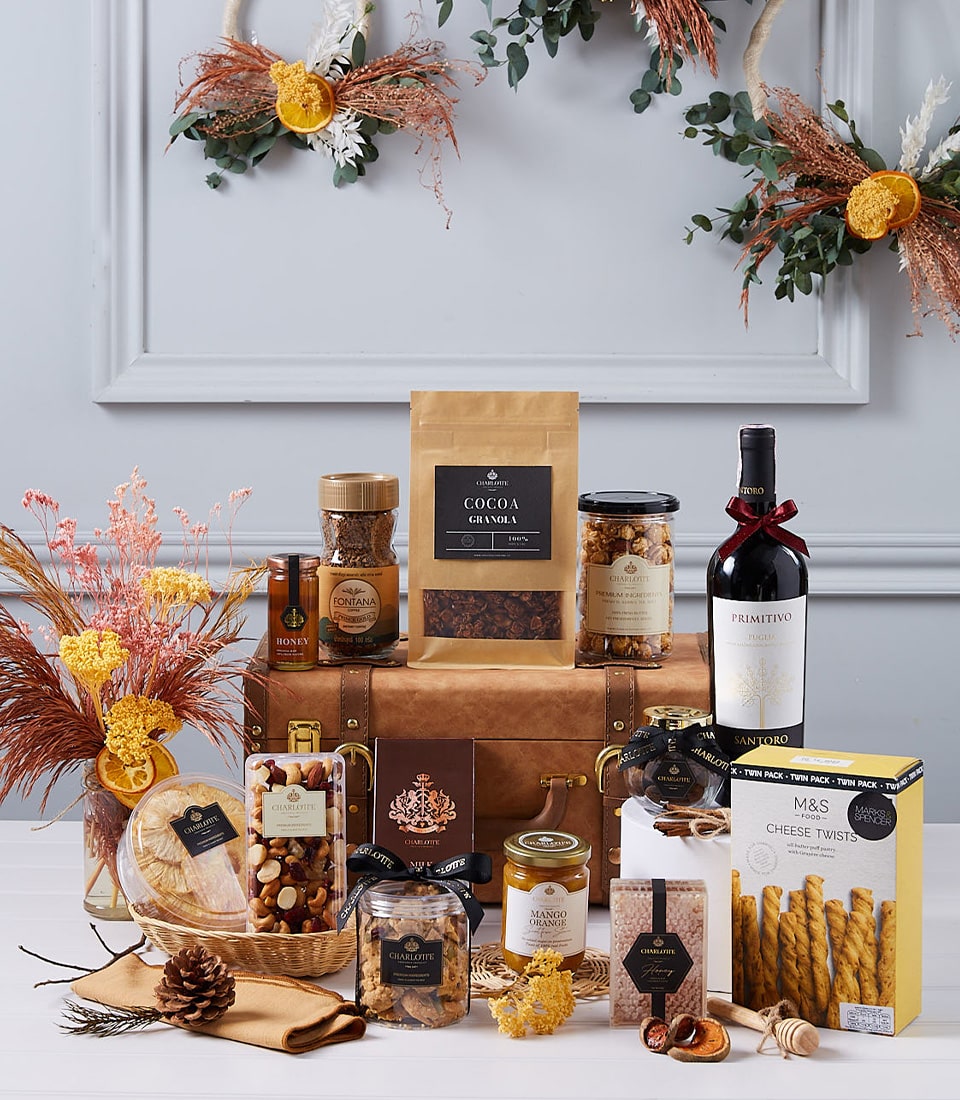A luxurious Basketeer gift hamper is elegantly displayed on a refined table, brimming with an exquisite selection of gourmet delights. A fine bottle of wine takes center stage amidst an array of indulgent snacks, artisanal chocolates, premium nuts, and delectable biscuits. The scene is beautifully framed by stunning autumnal floral decorations that adorn a light gray wall in the background, enhancing the inviting and seasonal ambiance. A ribbon adorned with the Basketeer logo adds a touch of sophistication to this opulent display.