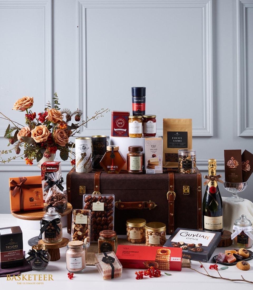 An opulent gift hamper by Basketeer elegantly presented atop and around a grand chest. The assortment includes fine wines, artisanal chocolates, golden honey, premium nuts, sumptuous jams, and delectable cookies. This luxurious selection is gracefully adorned with a lush floral arrangement and vibrant red berries. Some items are nestled in beautifully crafted boxes showcasing the Basketeer logo, enhancing the sophistication of this inviting scene.