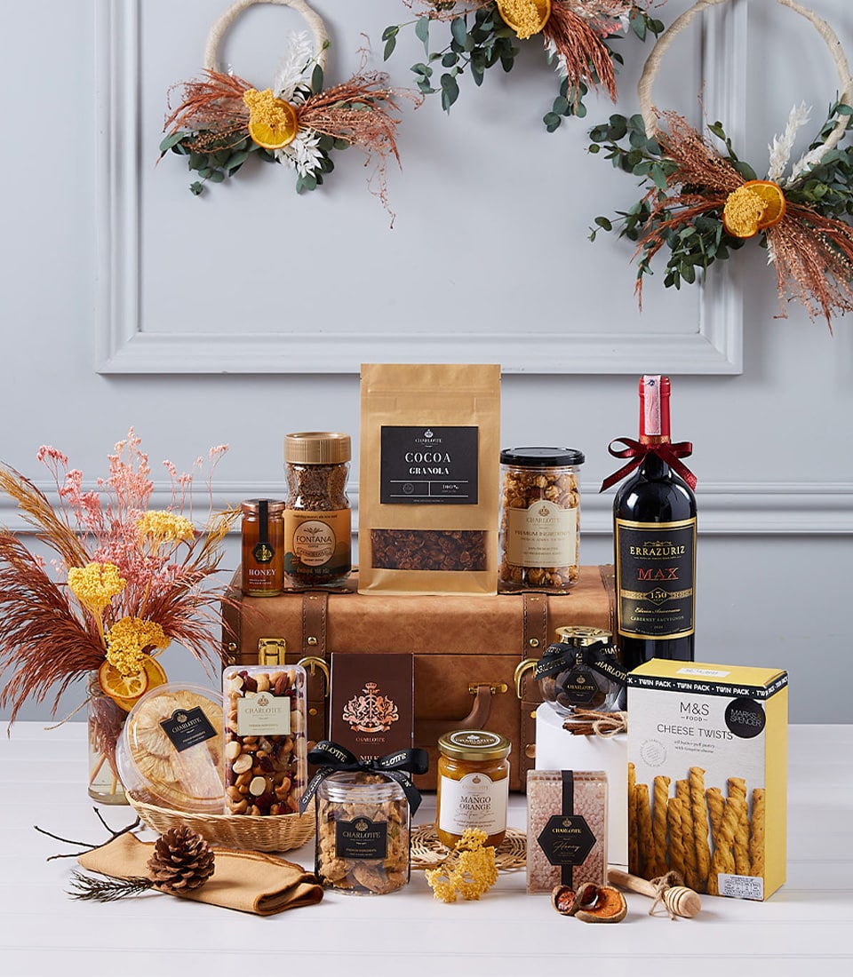 An opulent ensemble of gourmet indulgences presented by Basketeer, featuring an elegantly wrapped bottle of fine red wine, artisanal cocoa granola, savory cheese twists, luscious honey, premium nuts, delectable spreads, and rich chocolate. This luxurious hamper is meticulously arranged amidst charming decorative wreaths and dried floral assortments on a polished wooden surface. The signature Basketeer logo graces the ribbon adorning the gift box, enhancing the scene with a touch of refined sophistication.