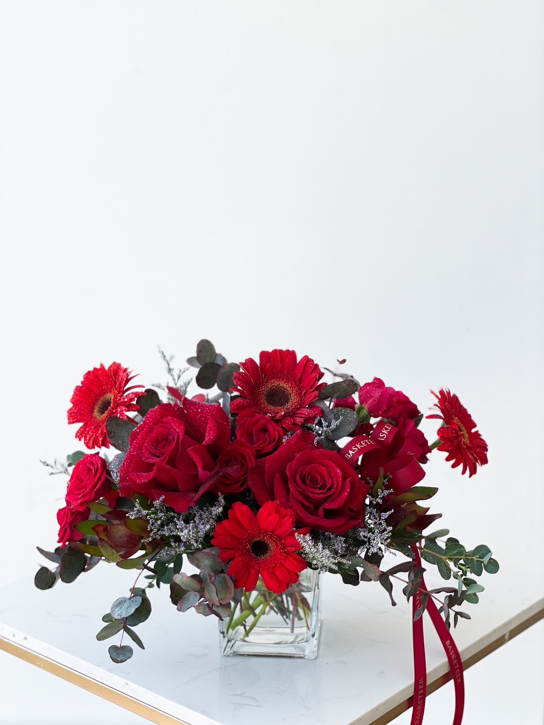 A rectangular glass vase holds an arrangement of vibrant red roses, gerbera daisies, and silver-green eucalyptus leaves. A red ribbon is draped artistically around the flowers, all set against a clean, white background.