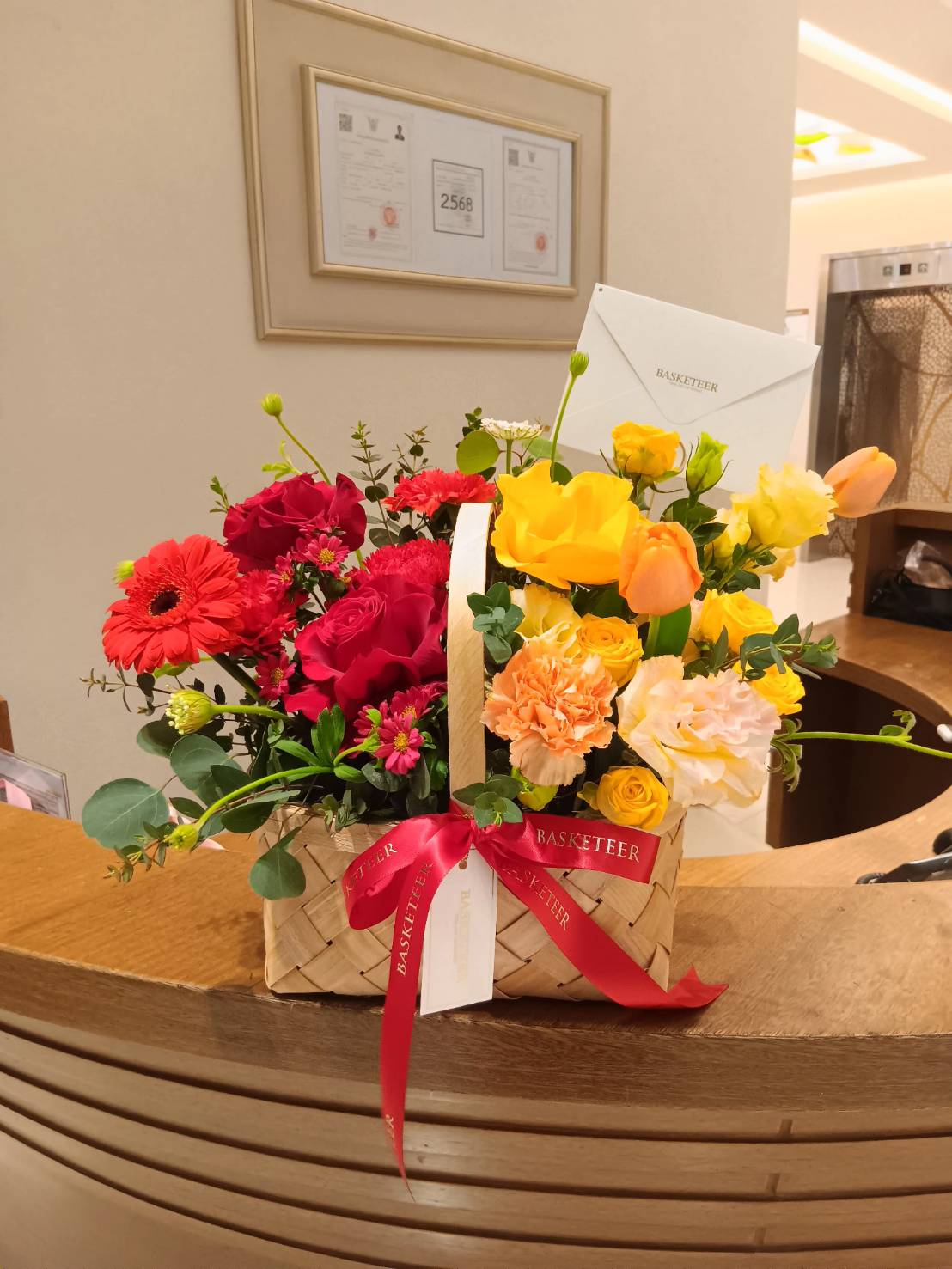 A stunning floral arrangement in a woven basket, featuring roses, carnations, and gerberas in warm sunset hues of red, yellow, and orange.