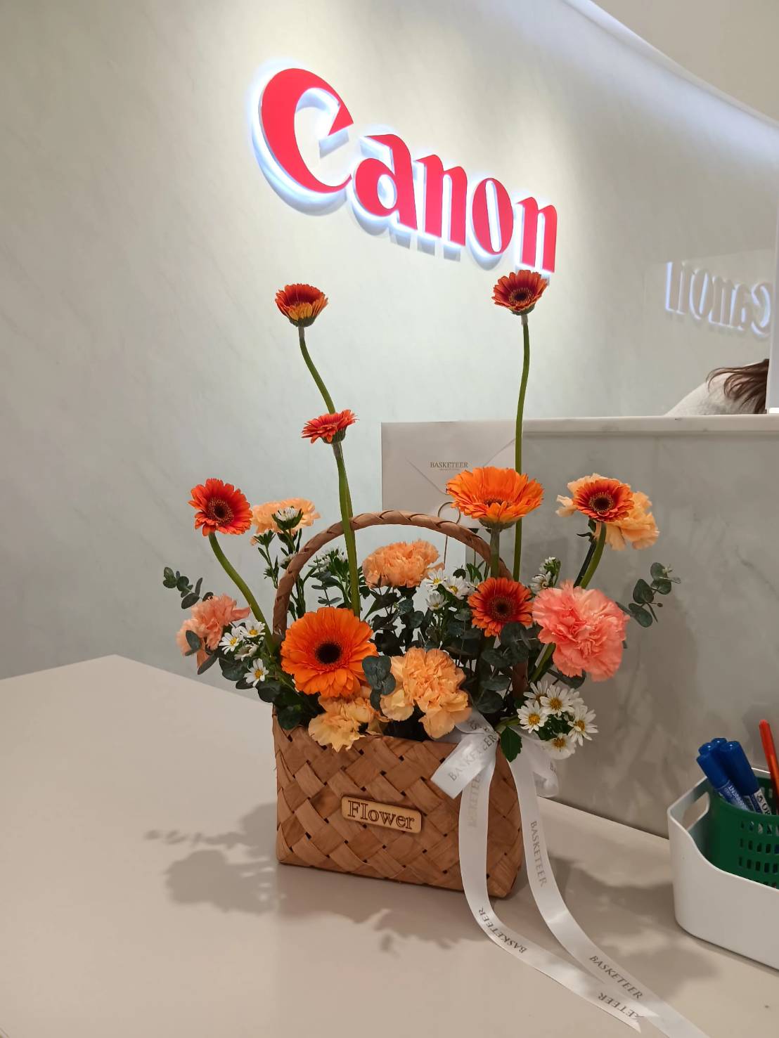 A handwoven basket filled with fresh orange gerbera daisies, carnations, and white flowers, arranged elegantly with a signature ribbon.