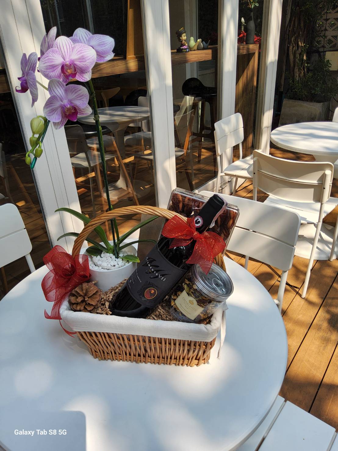 A luxurious wicker gift basket featuring a blooming purple orchid, a premium red wine bottle, and a jar of gourmet snacks, adorned with festive red ribbons.