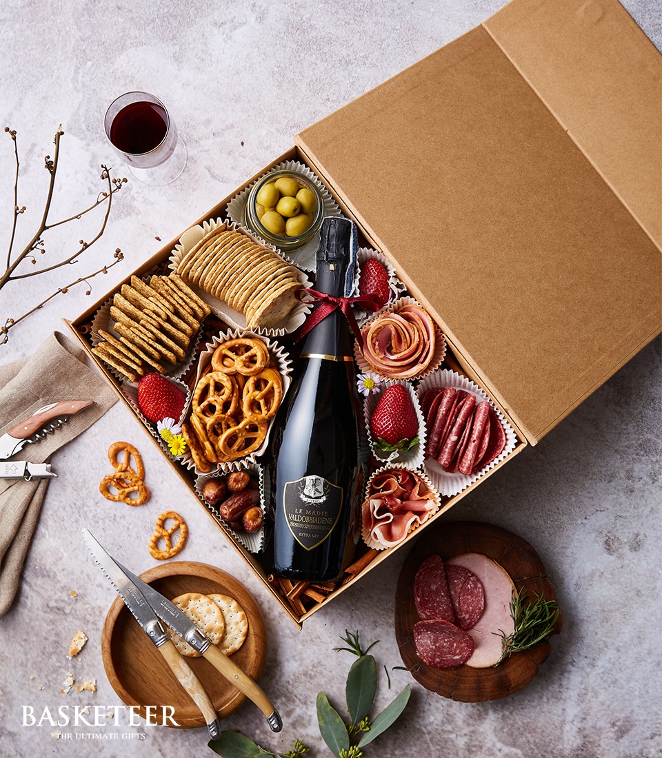 A luxurious Basketeer gift box showcasing a bottle of exquisite sparkling wine, an assortment of gourmet crackers, elegantly twisted pretzels, succulent green olives, fresh strawberries perfectly ripe for indulgence, artisan sliced cured meats, a selection of premium nuts, and a small block of rich cheese. The scene is further elevated with a glass of fine red wine nearby, accompanied by a refined bowl filled with delicate meat slices. A delectable cheese block adorned with an elegant knife awaits alongside an arrangement of crunchy cheese crackers. Finishing touches include the Basketeer logo ribbon gracefully draped over the box, emphasizing its opulence and inviting allure.