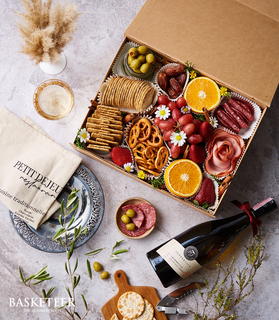 A luxurious charcuterie gift box by Basketeer, featuring an exquisite assortment of gourmet snacks. The elegantly arranged selection includes artisanal crackers, aged cheese, plump grapes, fresh strawberries, savory pretzels, briny olives, delectable slices of salami, and vibrant orange segments. The open box exudes sophistication and is accompanied by a refined setup: a premium wine bottle ready to be uncorked, a delicate plate holding extra crackers and olives alongside meticulously cut salami slices, and a beautifully folded cloth napkin embroidered with 