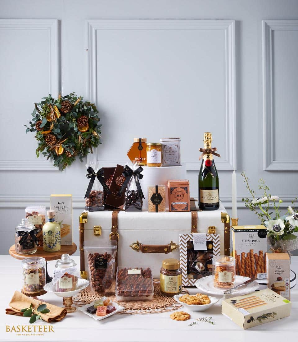 A lavishly arranged display showcases an elegant large white chest, brimming with a curated selection of gourmet delights. Surrounding the chest are exquisite bottles of fine wine, artisanal preserves, delectable pastries, luxurious chocolates, and savory snacks that tempt the senses. A festive wreath graces the backdrop with seasonal charm. The distinguished 