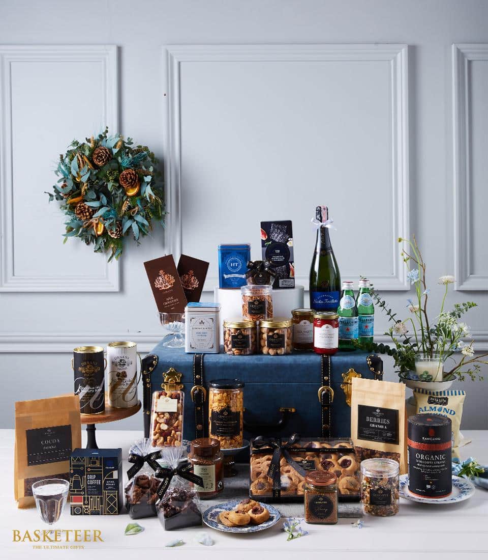 A luxurious gift hamper curated with finesse and elegance by Basketeer is artfully arranged on and around a sophisticated blue trunk. The assortment includes a bottle of fine wine, exquisite snacks, delectable cookies, artisanal chocolates, and gourmet preserves. Each item exudes opulence and promises indulgence. Above this stylish display hangs a beautifully crafted wreath adorned with an elegant bouquet of flowers. The tasteful scene whispers an invitation to savor the extraordinary quality that defines Basketeer’s offerings.