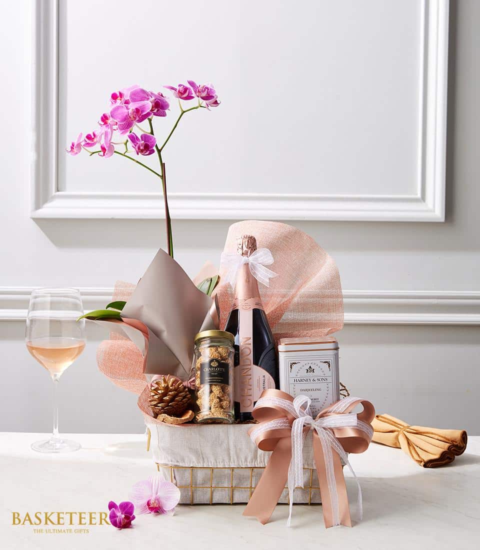 A lavish gift basket featuring a delicate pink orchid, an exquisite bottle of fine champagne, a luxurious candle, and a gourmet tin of snacks graces the elegantly set table. Complementing this decadent arrangement is a glass of rich Red Rose wine. The Bliss Basket is beautifully adorned with pink and white ribbon, exuding elegance and charm. Proudly displayed at the bottom left in sumptuous gold text is 