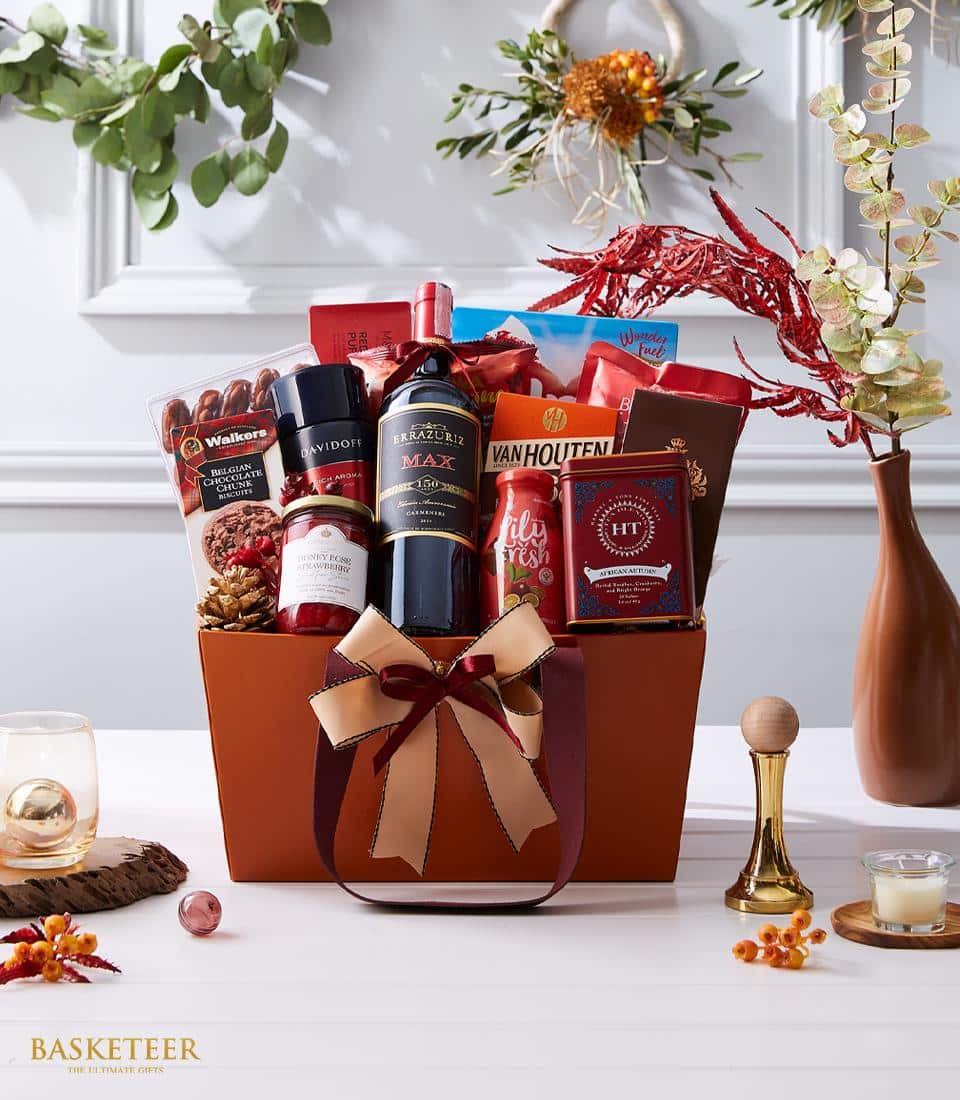 A festive gift basket adorned with a bow contains wine, chocolates, wafer rolls, nuts, and assorted holiday treats. The basket is surrounded by holiday decorations, including a candle, gold bell, and a small vase with dried flowers. The word 