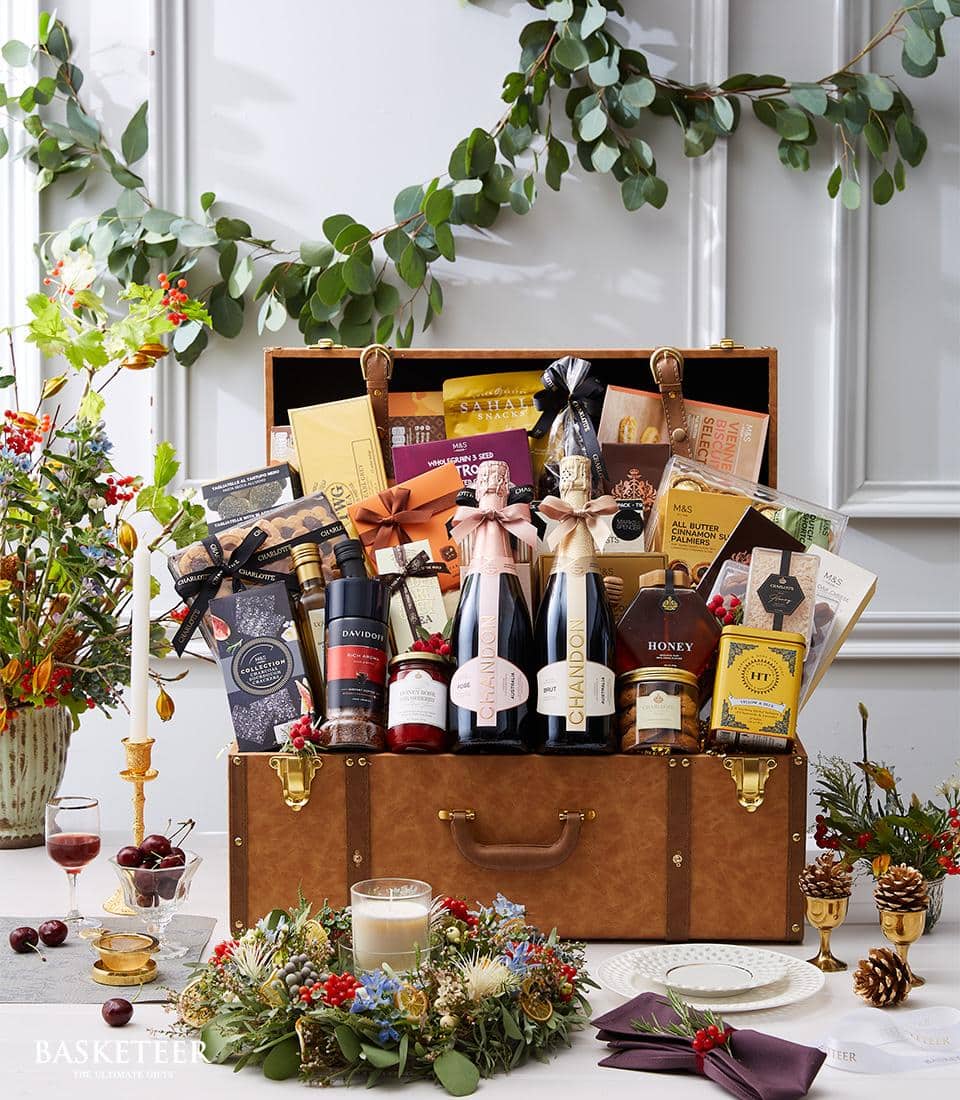 An opulently curated gift hamper nestled within an elegantly open brown suitcase, showcasing an exquisite selection of gourmet delights. Discover fine wine bottles, decadent chocolates, artisanal crackers, rich jams, and pure honey—all harmoniously arranged to tantalize the senses. The display is enhanced with luxurious festive greenery, delicate flowers, and shimmering candles on a beautifully adorned table. A ribbon adorned with the Basketeer logo bespeaks quality and sophistication, making this ensemble a truly memorable indulgence.