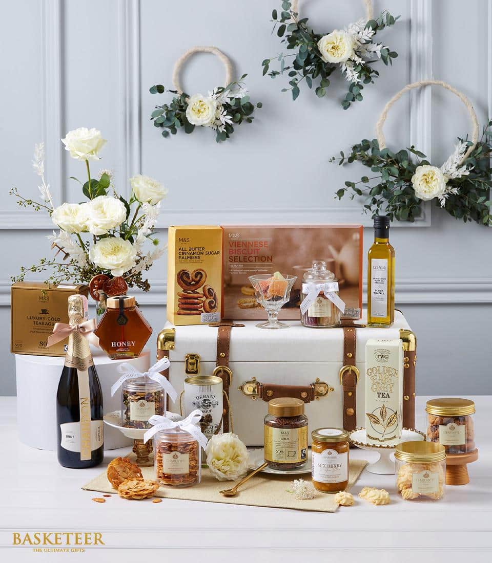 A luxurious Basketeer gift hamper brimming with an exquisite selection of gourmet delicacies is set against an opulent backdrop adorned with elegant floral arrangements. Featuring premium champagne, artisanal honey, lush preserves, decadent chocolates, and other indulgent treats, each item is meticulously arranged around a pristine white and gold vintage-style trunk. A sophisticated Basketeer ribbon gracefully drapes over the scene, promising an experience of true luxury and refinement.