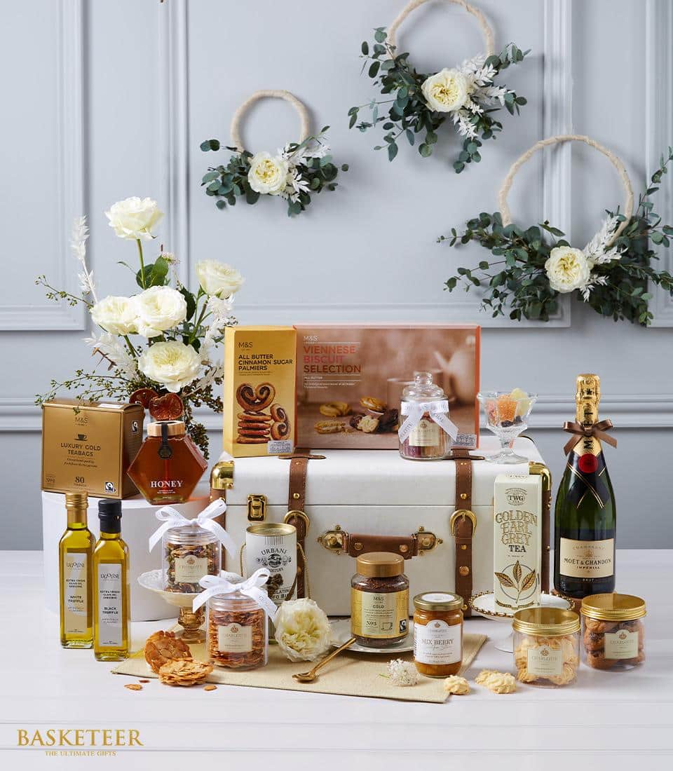 A luxurious Basketeer gift hamper elegantly displayed on a sophisticated table setting. The artistic arrangement features an array of gourmet delights including opulent tea, golden honey, premium olive oil, artisanal cookies, a rich jar of pasta sauce, decadent chocolates, and an exquisite bottle of fine wine. Delicate decorative flowers and lush greenery serve as a charming backdrop. A tasteful white velvet ribbon adorned with the Basketeer logo enhances the presentation, embodying both elegance and indulgence.
