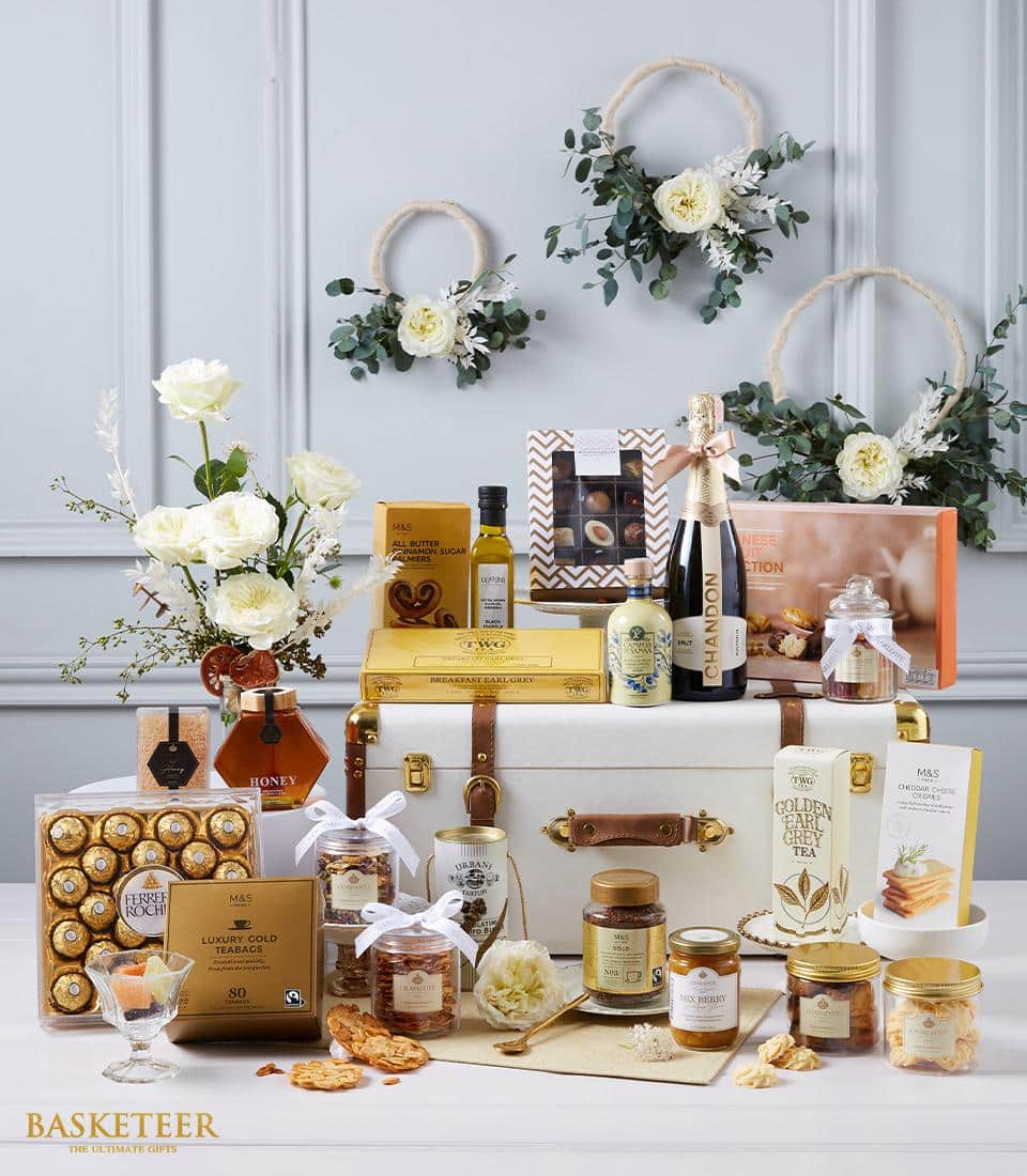 A lavish gift display curated by Basketeer, showcasing an elegant white chest encircled by an opulent selection of gourmet delicacies. The assortment features exquisite honey, premium tea, decadent chocolates, rich olive oil, artisanal biscuits, and sumptuous jam. Surrounding the scene are sophisticated floral arrangements and lush greenery that enhance the inviting atmosphere. A chic ribbon branded with the Basketeer logo gracefully accents the presentation box.