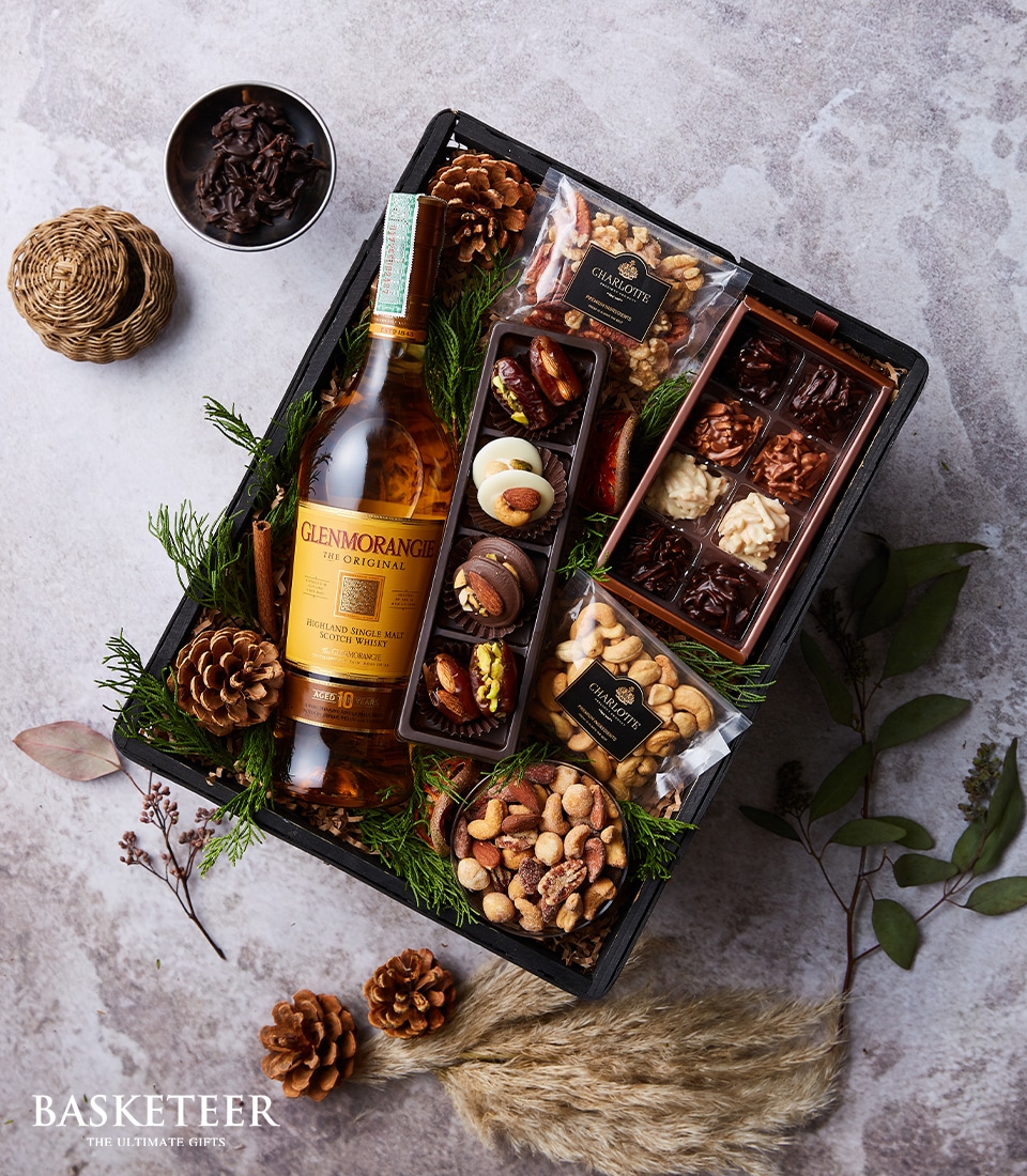 An exquisite and inviting gift basket adorned with the premium Glenmorangie Original whisky, elegantly nestled amid festive holiday greenery and charming pinecones. The assortment includes gourmet chocolates, delectable dried fruits, and a selection of fine nuts, all beautifully presented in a chic black tray from Basketeer. Luxuriously tied with a refined ribbon featuring the distinguished Basketeer logo, this gift is perfect for indulging in the season's celebrations.