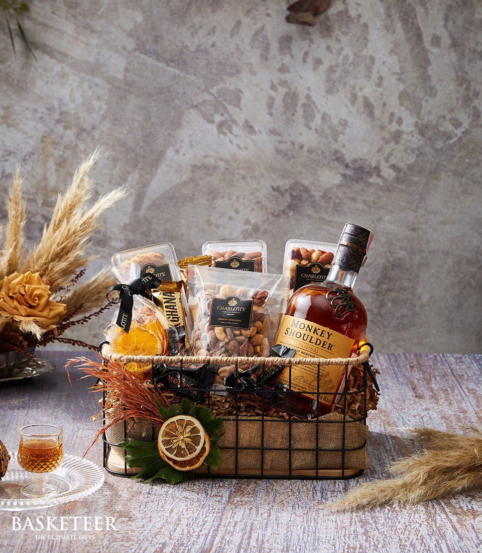 An exquisitely curated gift basket by Basketeer graces a refined table, showcasing a bottle of premium Monkey Shoulder whisky alongside an assortment of gourmet snacks. Artfully arranged, the ensemble includes elegantly packaged nuts, dried fruits, and other delectable treats. Decorative dried plants adorn the display, adding a touch of natural sophistication. A luxurious Basketeer ribbon subtly accents the presentation against a backdrop of rustic textured walls, inviting you to indulge in opulent relaxation.