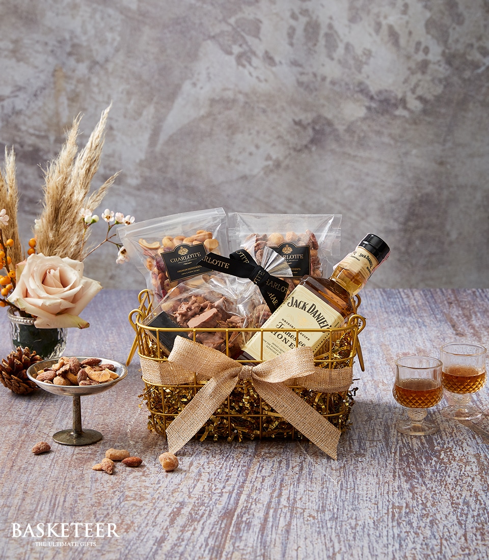A luxurious Basketeer gift basket rests elegantly on a rustic wooden table, brimming with an assortment of premium nuts, two exquisite small bottles of Jack Daniel's whiskey, and a decadent pack of gourmet chocolate. The basket is beautifully adorned with a burlap bow featuring the sophisticated Basketeer logo. Nearby, two refined whiskey glasses await indulgence beside a delicate peach rose and an arrangement of light brown dried flowers in a charming vase. This inviting scene exudes warmth and opulence, perfect for any discerning connoisseur.