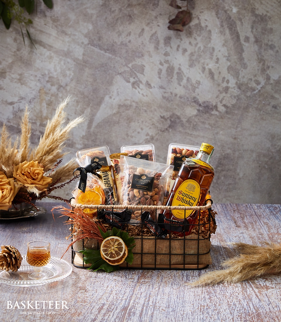 A luxurious gift basket from Basketeer showcasing Suntory whisky, elegantly paired with premium bags of assorted nuts and artistically arranged dried orange slices. Nestled amidst decorative dried plants, the basket sits on a rustic wooden table exuding charm. A ribbon adorned with the Basketeer logo complements the sophisticated design. Nearby, a glass of whisky awaits to be savored, set against a beautifully textured wall that enhances the opulent ambiance of the scene.