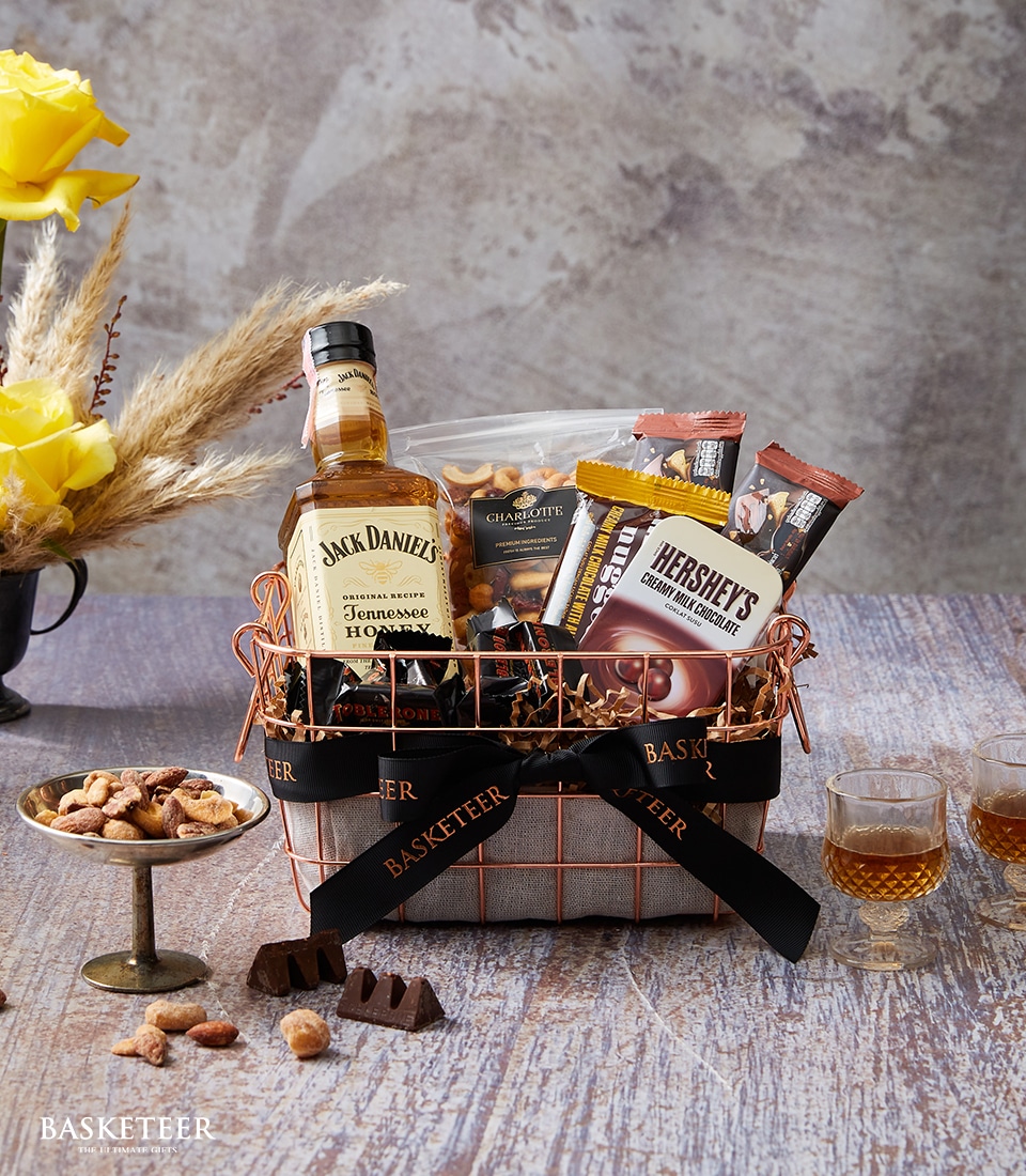 A meticulously curated Basketeer gift basket graces a polished wooden table, epitomizing both luxury and indulgence. Nestled within this elegant assortment is a bottle of Jack Daniel's whiskey, accompanied by decadent Hershey's chocolate bars and a gourmet selection of mixed nuts. Two refined glasses of whiskey stand ready to offer a taste of sophistication. The scene is further elevated by the presence of vibrant yellow flowers in a vase, adding a touch of organic beauty to the setting. The distinguished Basketeer logo adorns the ribbon wrapped around this exquisite collection, promising an unforgettable experience.