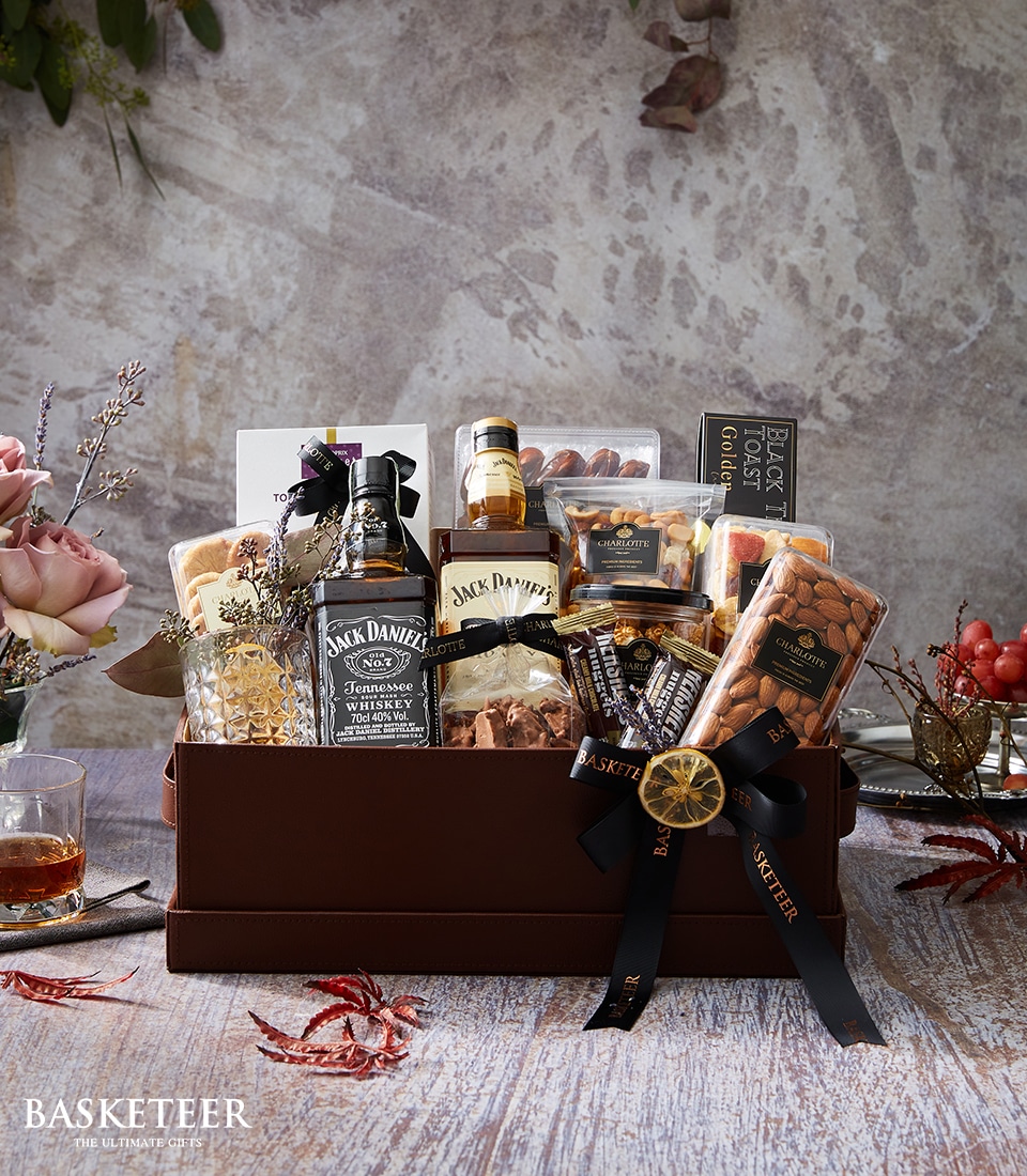 A lavish gift basket features the distinguished Jack Daniel's whiskey, an assortment of gourmet snacks, and decadent chocolates. These exquisite items are artfully arranged in a sophisticated brown leather box graced with the Basketeer logo and adorned with a chic black ribbon. The tableau is complemented by elegant flowers, autumn leaves, and a glistening glass of whiskey, creating an inviting scene of opulence and warmth.