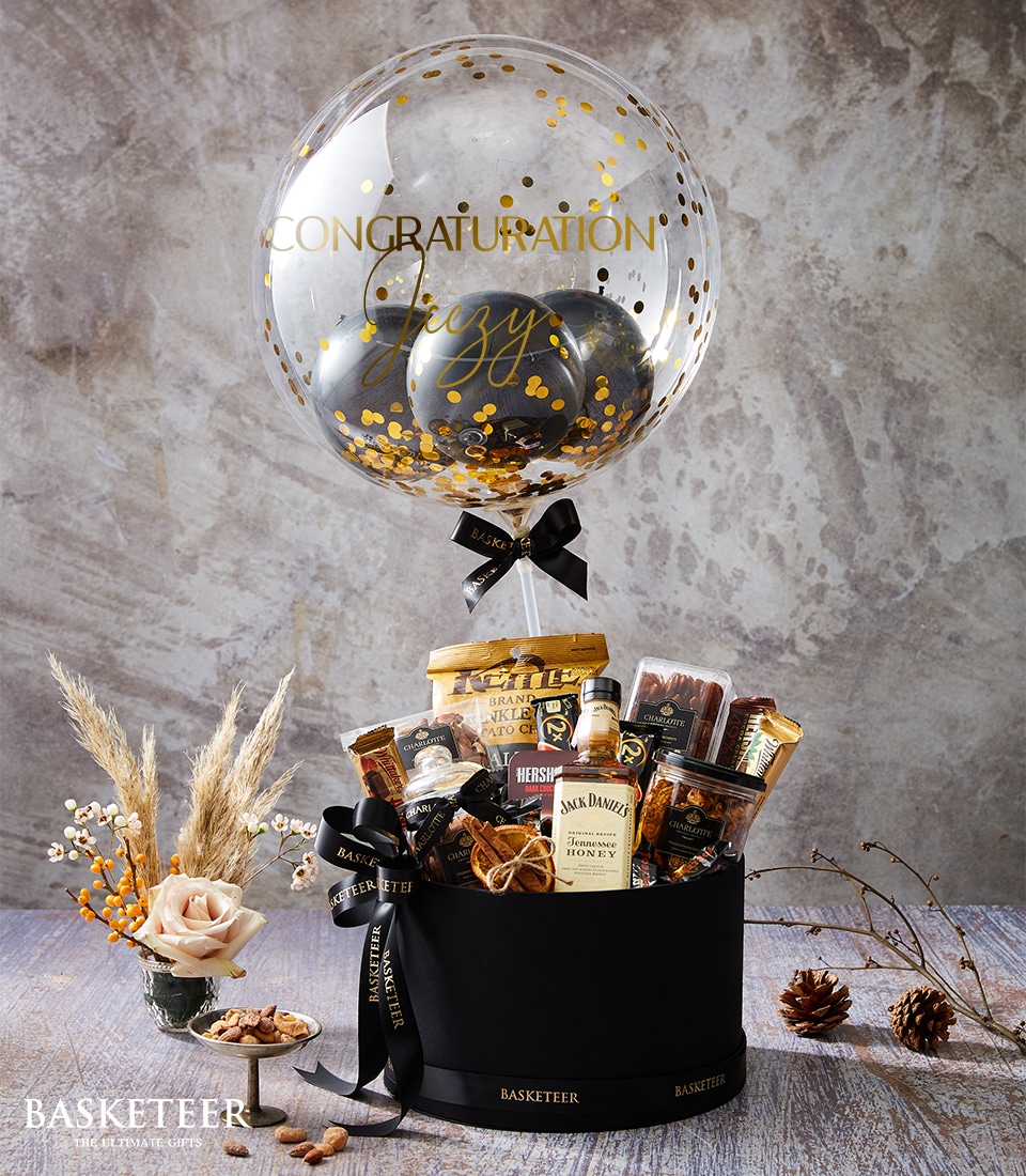 A lavish Basketeer gift basket graces a rustic wooden surface, exuding elegance and charm. This deluxe assortment features an array of premium treats including sumptuous chocolates, gourmet nuts, and a miniature bottle of Jack Daniel's nestled in fine packaging. Above this opulent display floats a clear balloon adorned with golden confetti, bearing a heartfelt congratulatory message. The scene is enhanced by tasteful decorative items, adding to the overall allure of this luxurious presentation. The ribbon and box proudly showcase the distinguished Basketeer logo, promising an experience of exceptional quality and indulgence.
