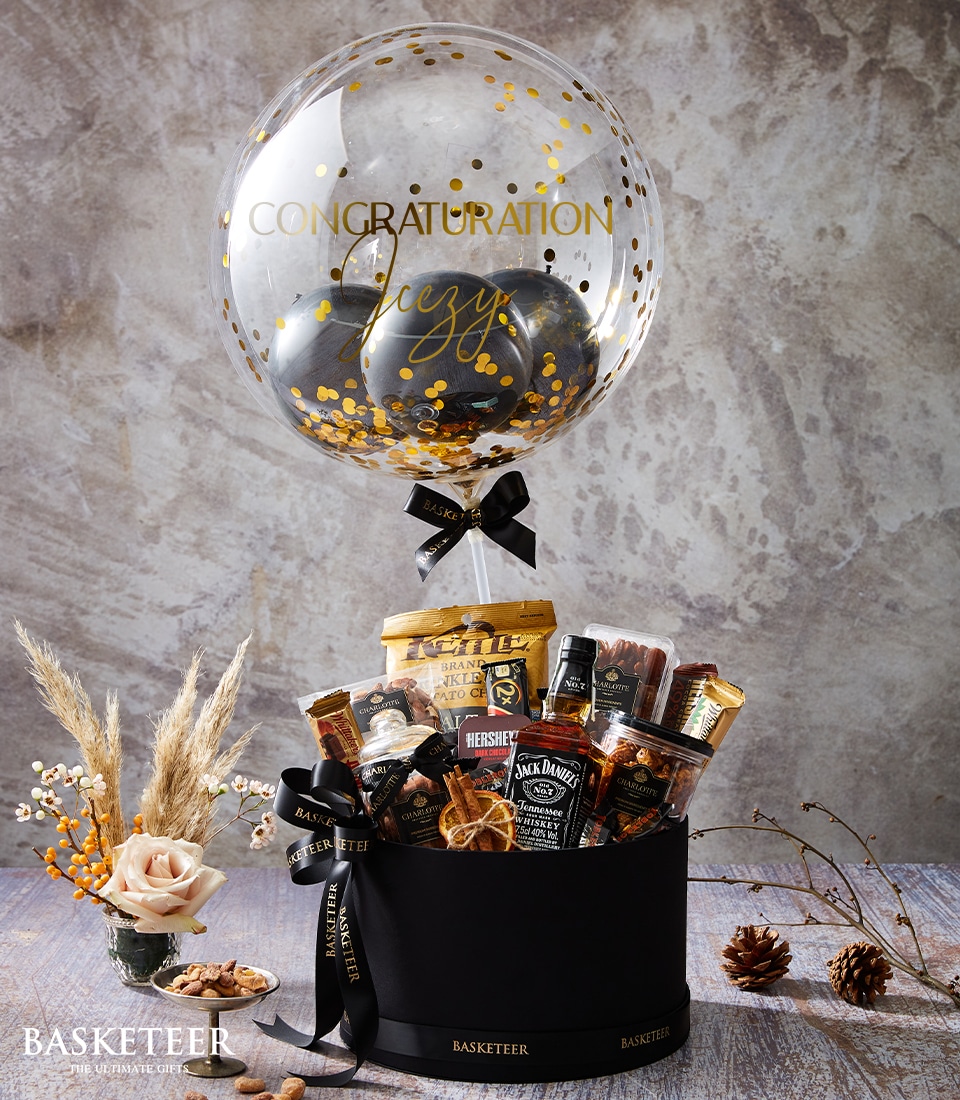 A sumptuous Basketeer gift basket graces an elegant table, exuding luxury and charm. A clear balloon filled with glistening gold confetti prominently floats above, showcasing the celebratory message 