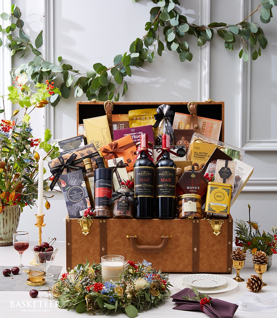 A luxurious gift hamper elegantly arranged within a polished, open brown suitcase, showcasing exquisite bottles of red wine alongside a curated selection of gourmet treats and decadent chocolates. The hamper is adorned with jars of premium honey and finished with festive decorations, exuding a sense of opulence. Nestled around the suitcase are enchanting wreaths, a softly glowing candle, an impeccably wrapped gift box featuring the distinguished Basketeer logo, and two sophisticated wine glasses brimming with rich red wine.