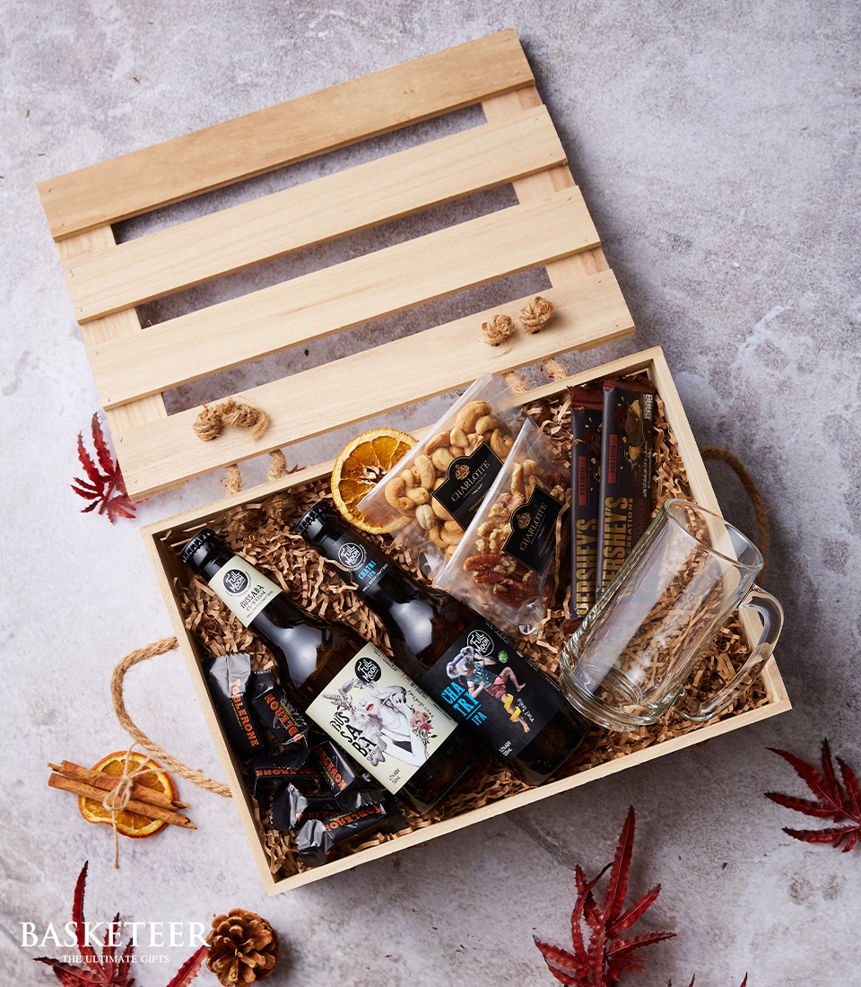 A luxurious wooden gift box, elegantly adorned with the prestigious Basketeer logo, is open on a light-colored surface embellished with scattered autumn leaves. Inside, an enticing collection awaits: artisanal craft beer bottles paired flawlessly with a crystal-clear beer mug. Also nestled within are gourmet snacks including savory nuts and premium beef jerky, alongside beautifully dried orange slices and decadent wrapped chocolates. The scene exudes an inviting ambiance of indulgence and warmth, perfect for savoring the finer moments.